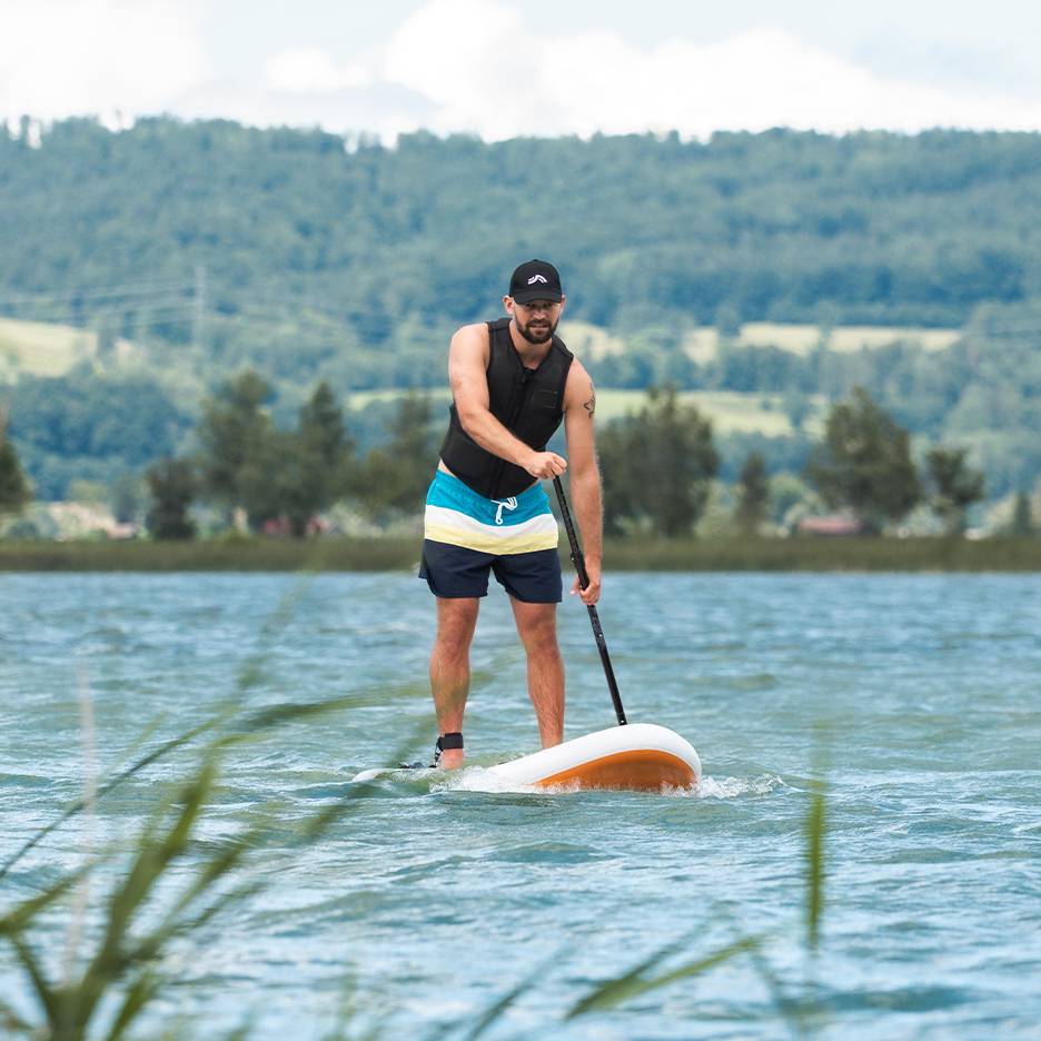 Άνδρας κάνει stand-up paddle σε λίμνη, με σωσίβιο και μαγιό.