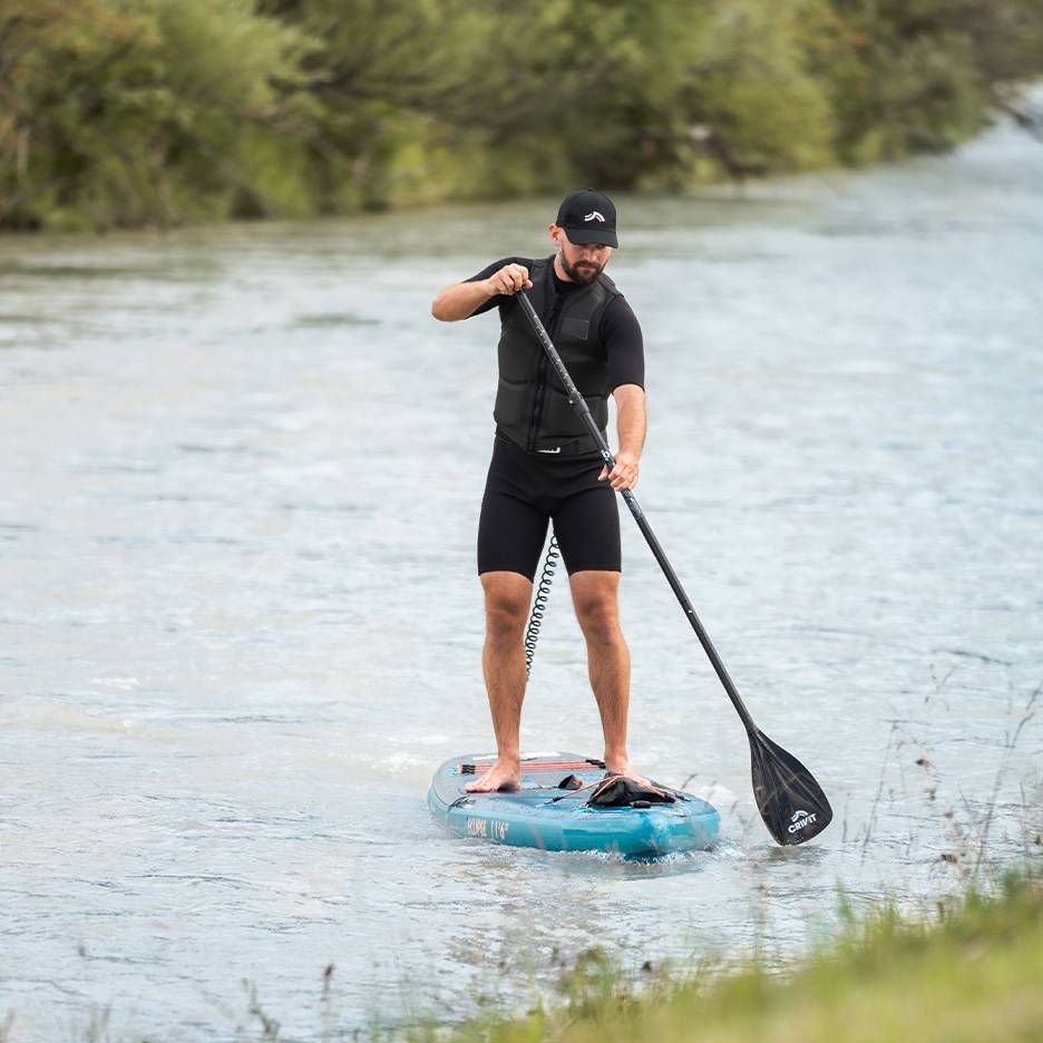 Άνδρας κάνει paddleboard σε ποτάμι, φορώντας μαύρη στολή, σωσίβιο και καπέλο.