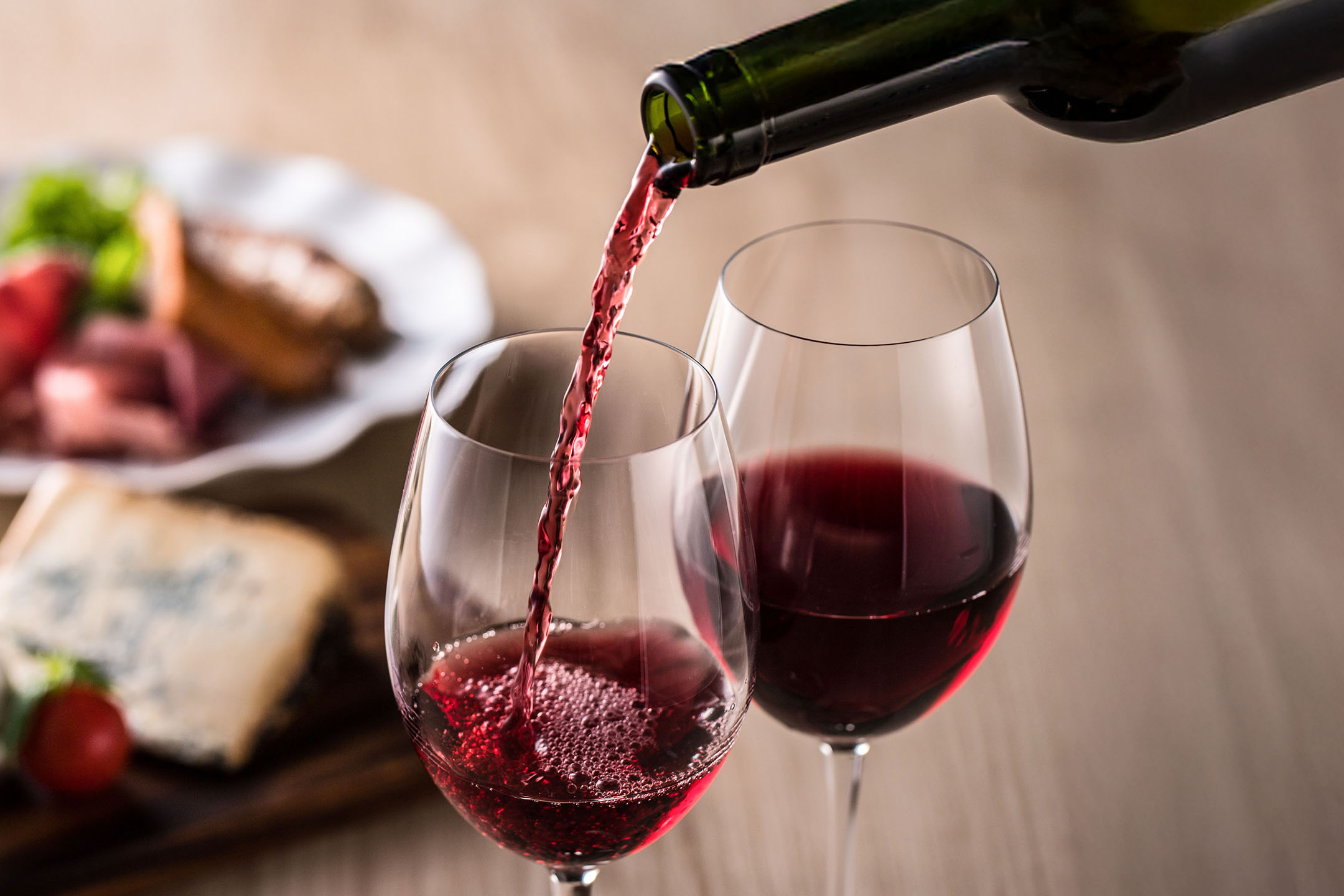 Κόκκινο κρασί χύνεται σε ποτήρι, με ορεκτικά στο παρασκήνιο.