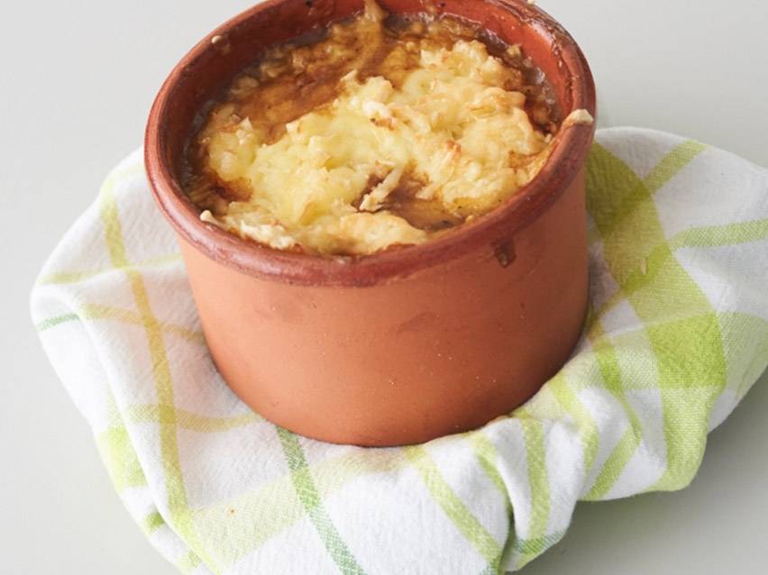 Πήλινο σκεύος με λιωμένο τυρί και καραμελωμένα κρεμμύδια, πάνω σε πετσέτα κουζίνας.