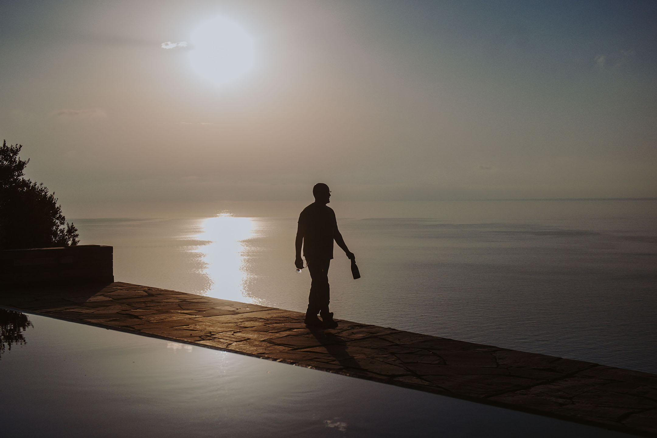 Σιλουέτα ατόμου με μπουκάλι και ποτήρι δίπλα σε πισίνα υπερχείλισης στο ηλιοβασίλεμα