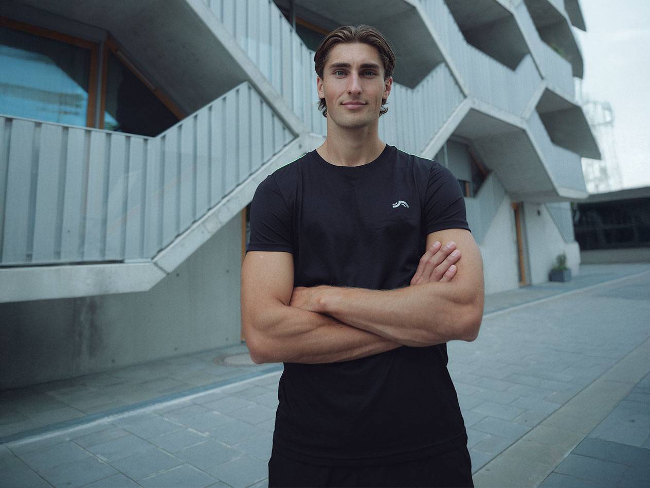 Άνδρας με μαύρο μπλουζάκι με λογότυπο και μαύρο σορτς, στέκεται σε εξωτερικό χώρο με σταυρωμένα χέρια.