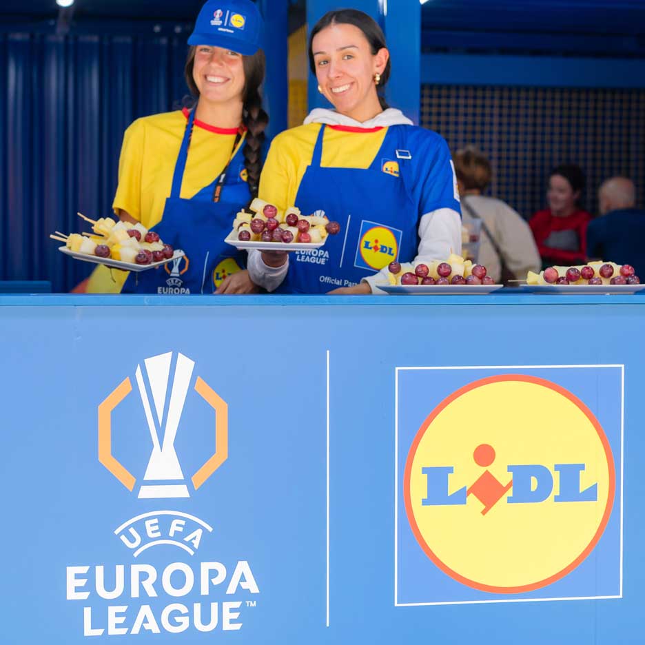 Δύο χαμογελαστές γυναίκες με ποδιές Lidl προσφέρουν φρούτα σε ξυλάκια σε εκδήλωση του UEFA Europa League.