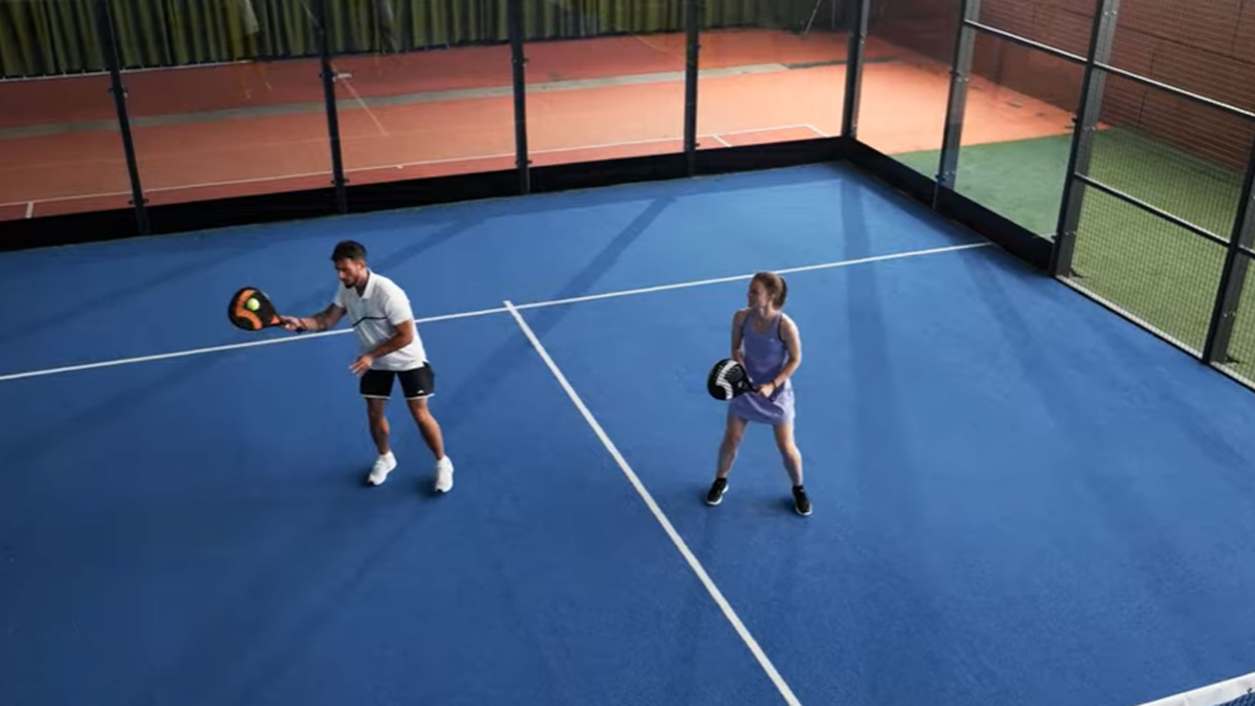 Άνδρας και γυναίκα παίζουν padel σε μπλε γήπεδο με ρακέτες και μπάλα