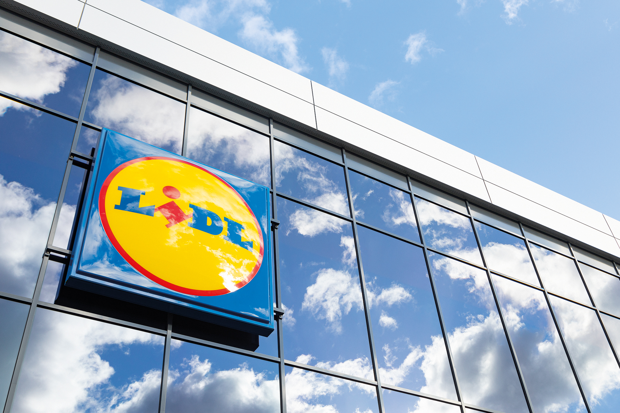 Πινακίδα Lidl σε γυάλινη πρόσοψη κτιρίου, με αντανάκλαση ουρανού και σύννεφων.