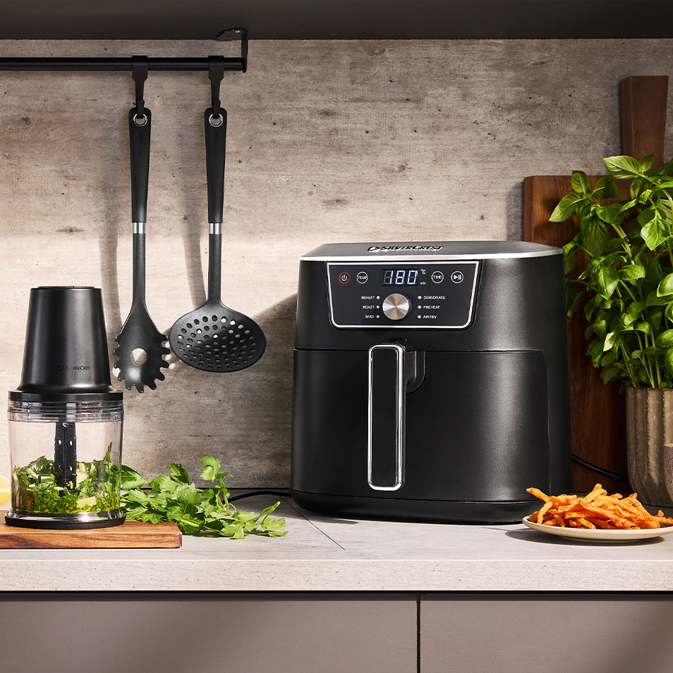 Φριτέζα αέρος SilverCrest και κόφτης τροφίμων σε πάγκο κουζίνας με φρέσκα βότανα και τηγανητές γλυκοπατάτες.