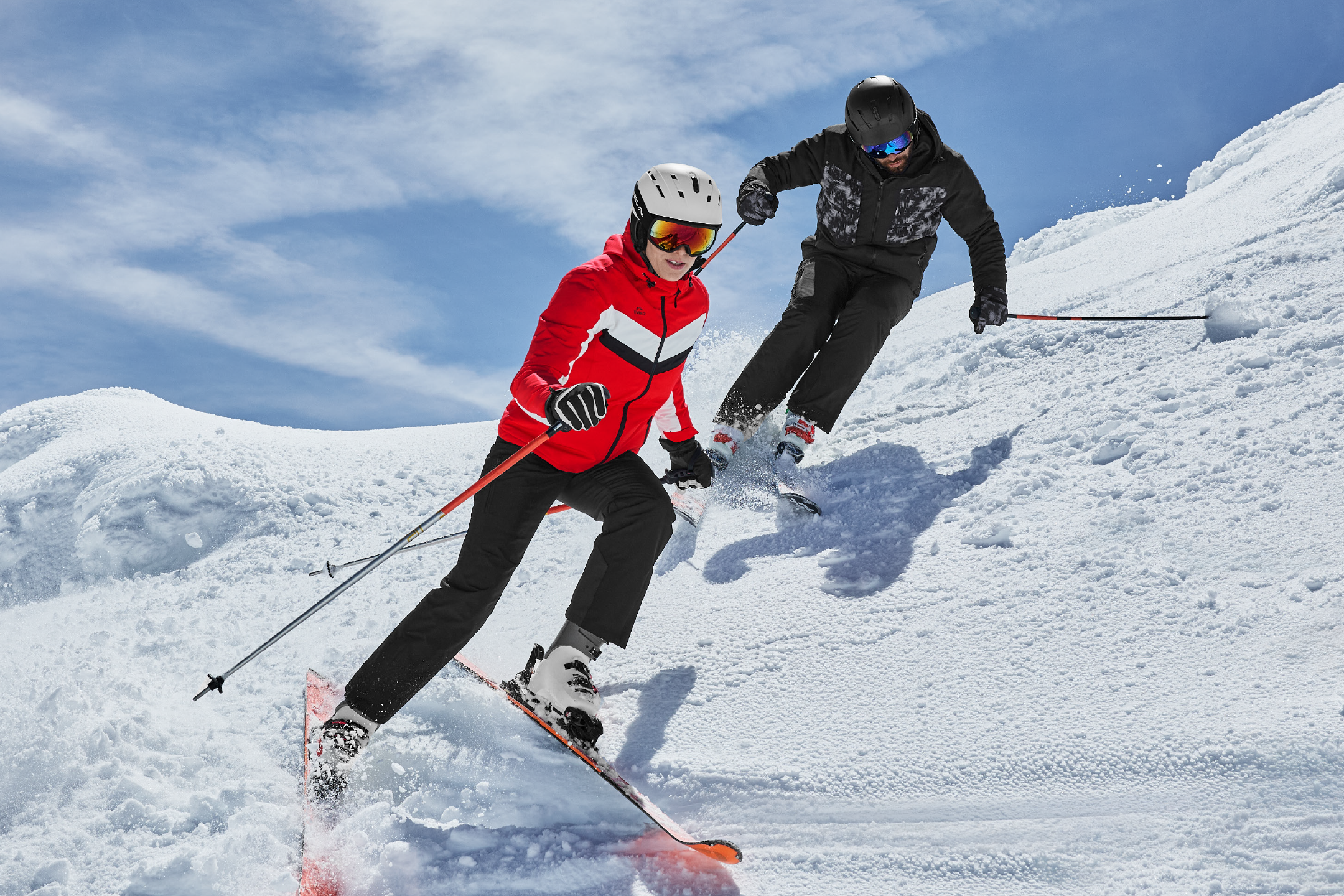 Δύο σκιέρ με κράνη και γυαλιά του σκι κατεβαίνουν μια χιονισμένη πλαγιά.