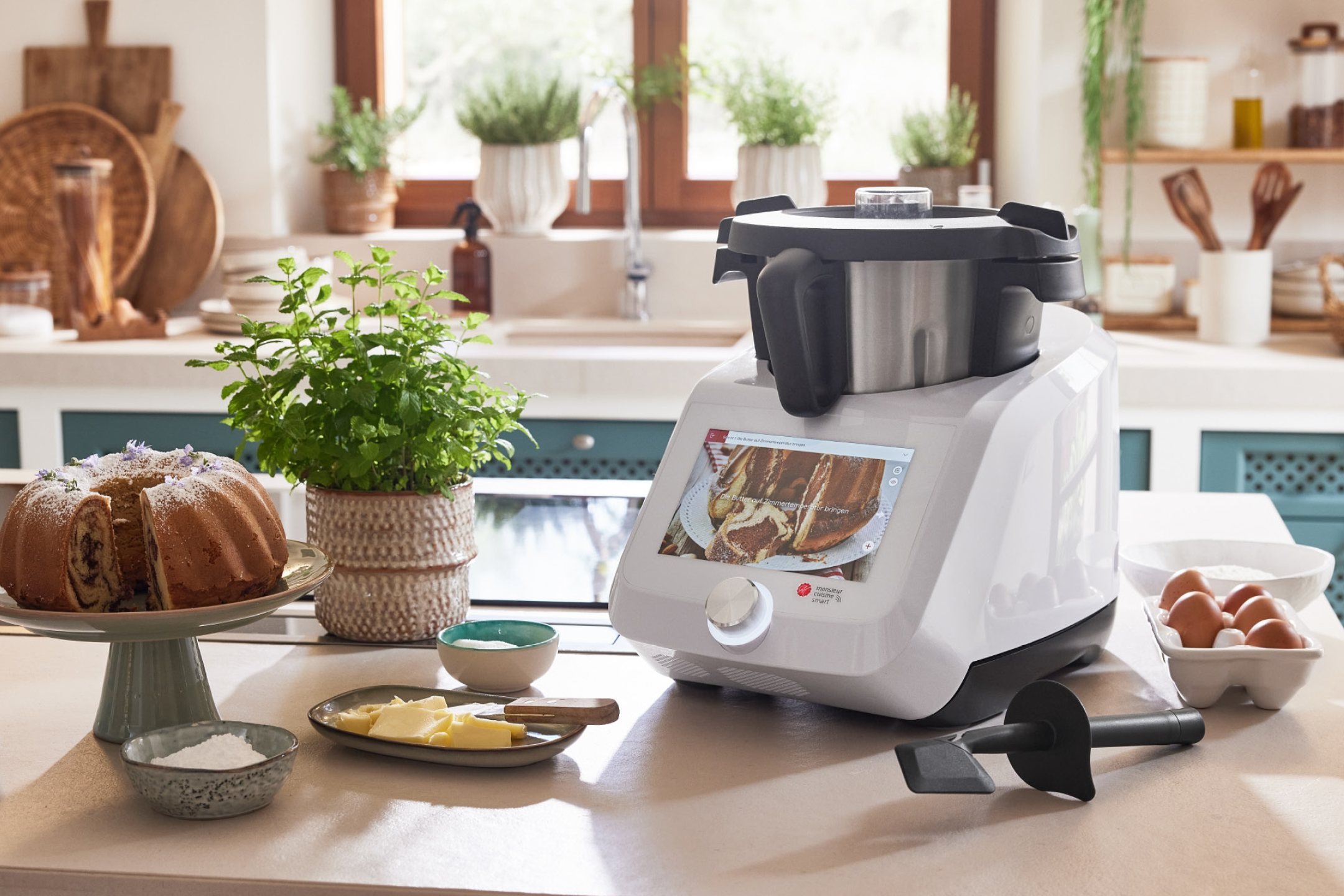 Κουζινομηχανή Monsieur Cuisine Smart σε πάγκο κουζίνας με κέικ, βούτυρο και αυγά.