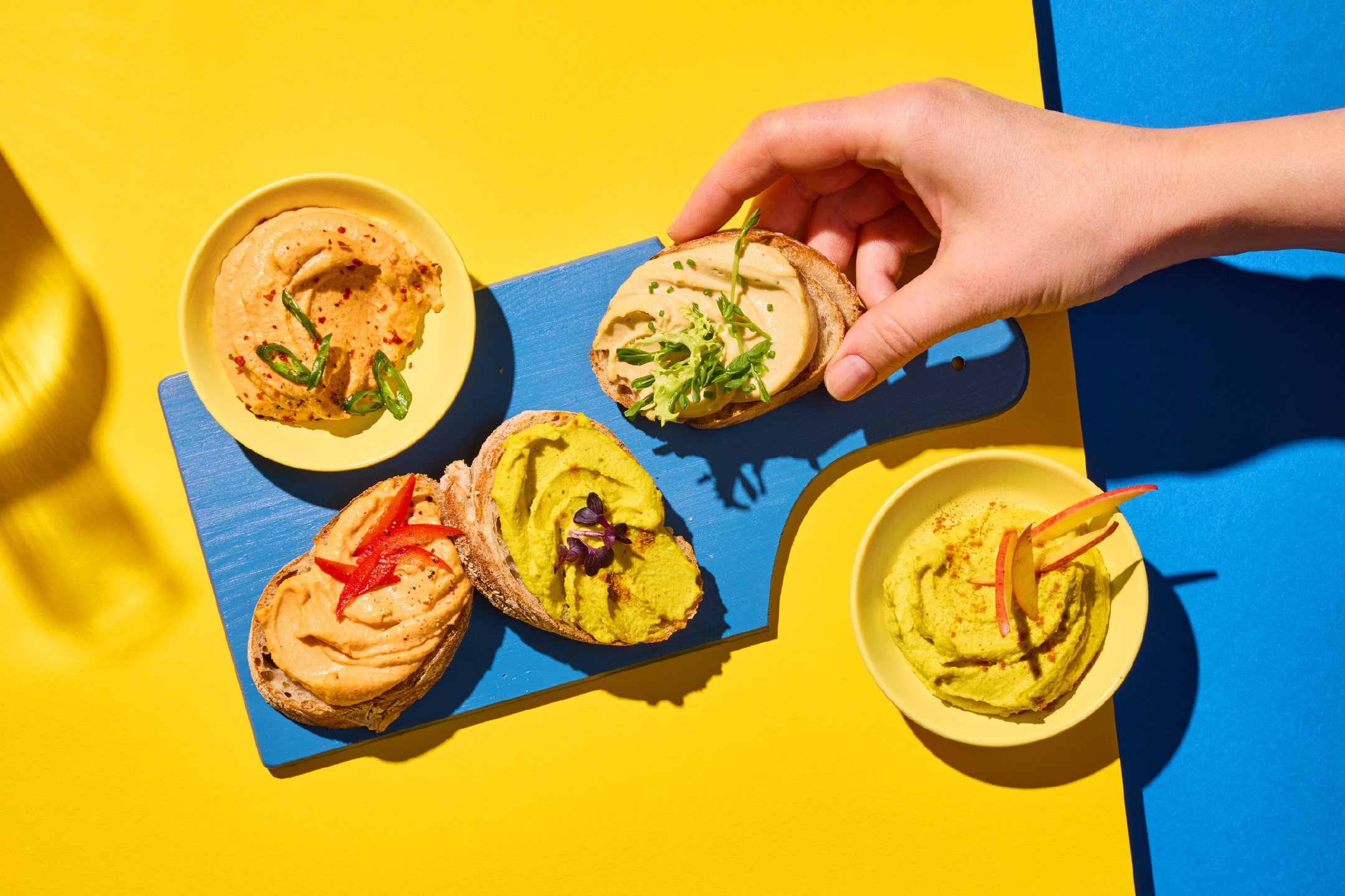 Χούμους σε μπολ και σε φρυγανιές με διάφορες γαρνιτούρες, σε κίτρινο και μπλε φόντο.