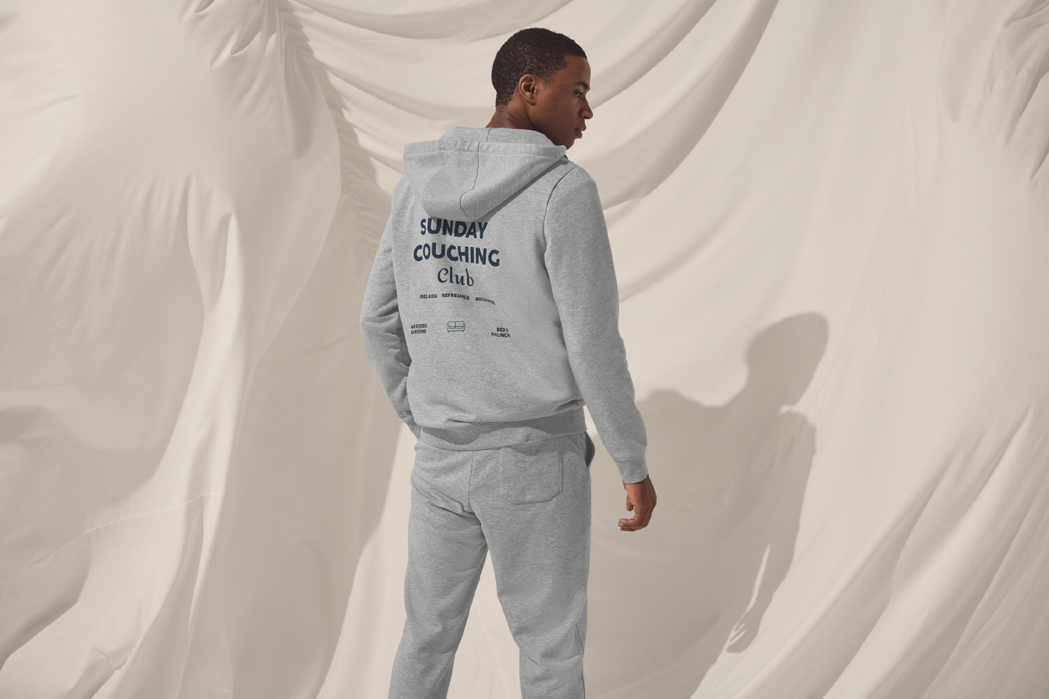 Άνδρας με γκρι φούτερ και παντελόνι φόρμας με την επιγραφή 'SUNDAY COUCHING Club' στην πλάτη.