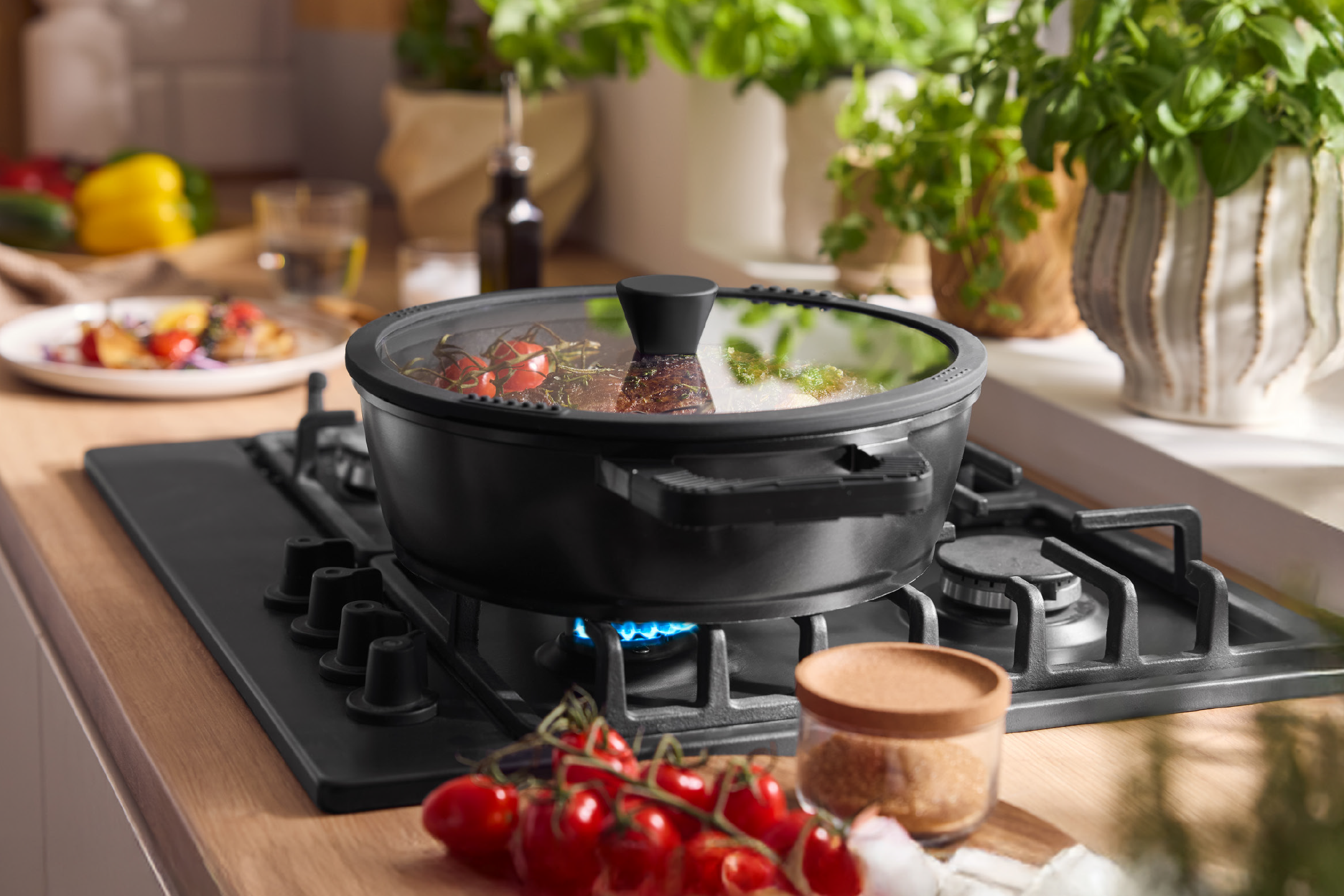Μαύρη κατσαρόλα με γυάλινο καπάκι σε εστία αερίου, με ντοματίνια και βότανα στο παρασκήνιο.