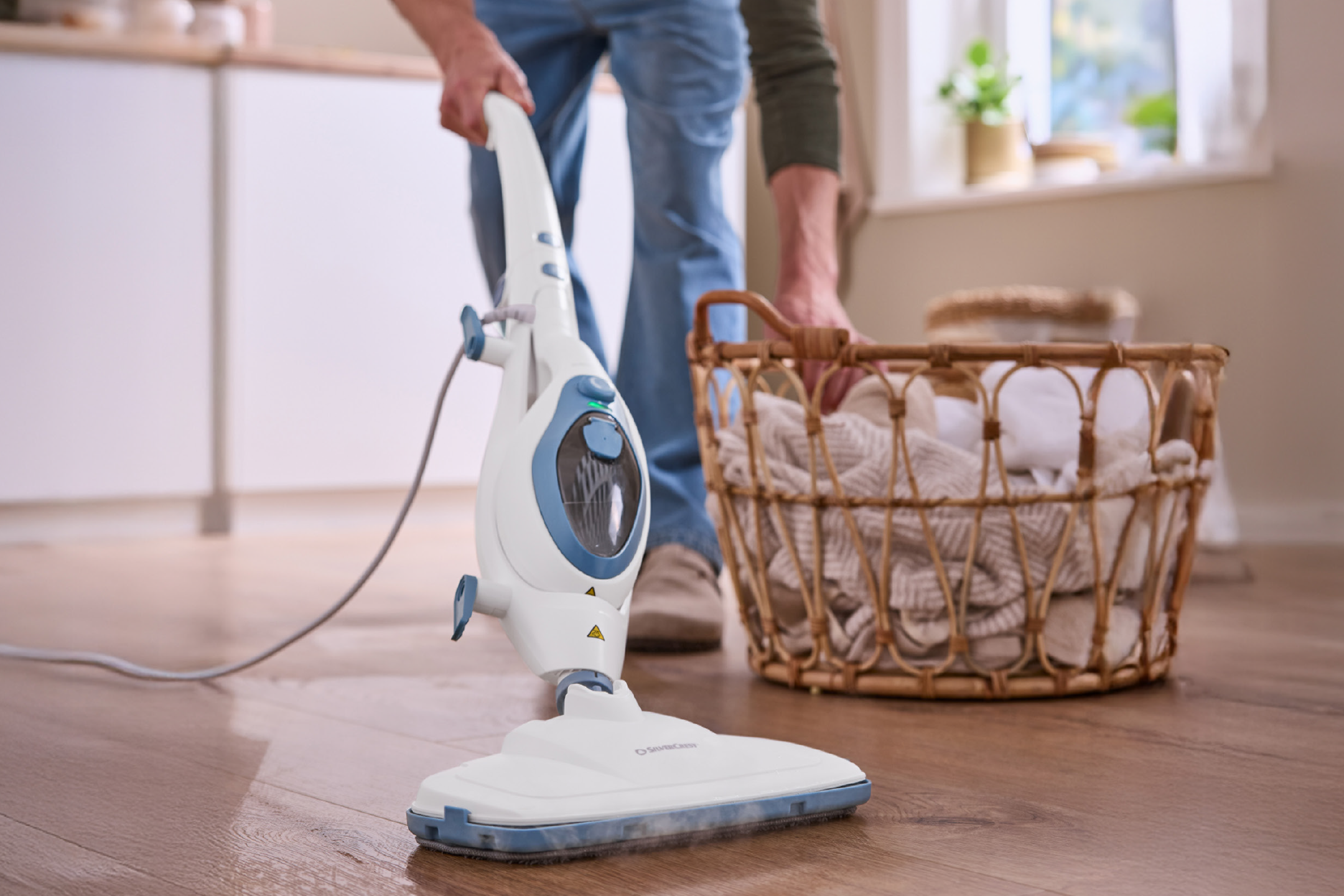 Άνδρας καθαρίζει ξύλινο πάτωμα με ατμοκαθαριστή SilverCrest, δίπλα σε καλάθι με ρούχα.