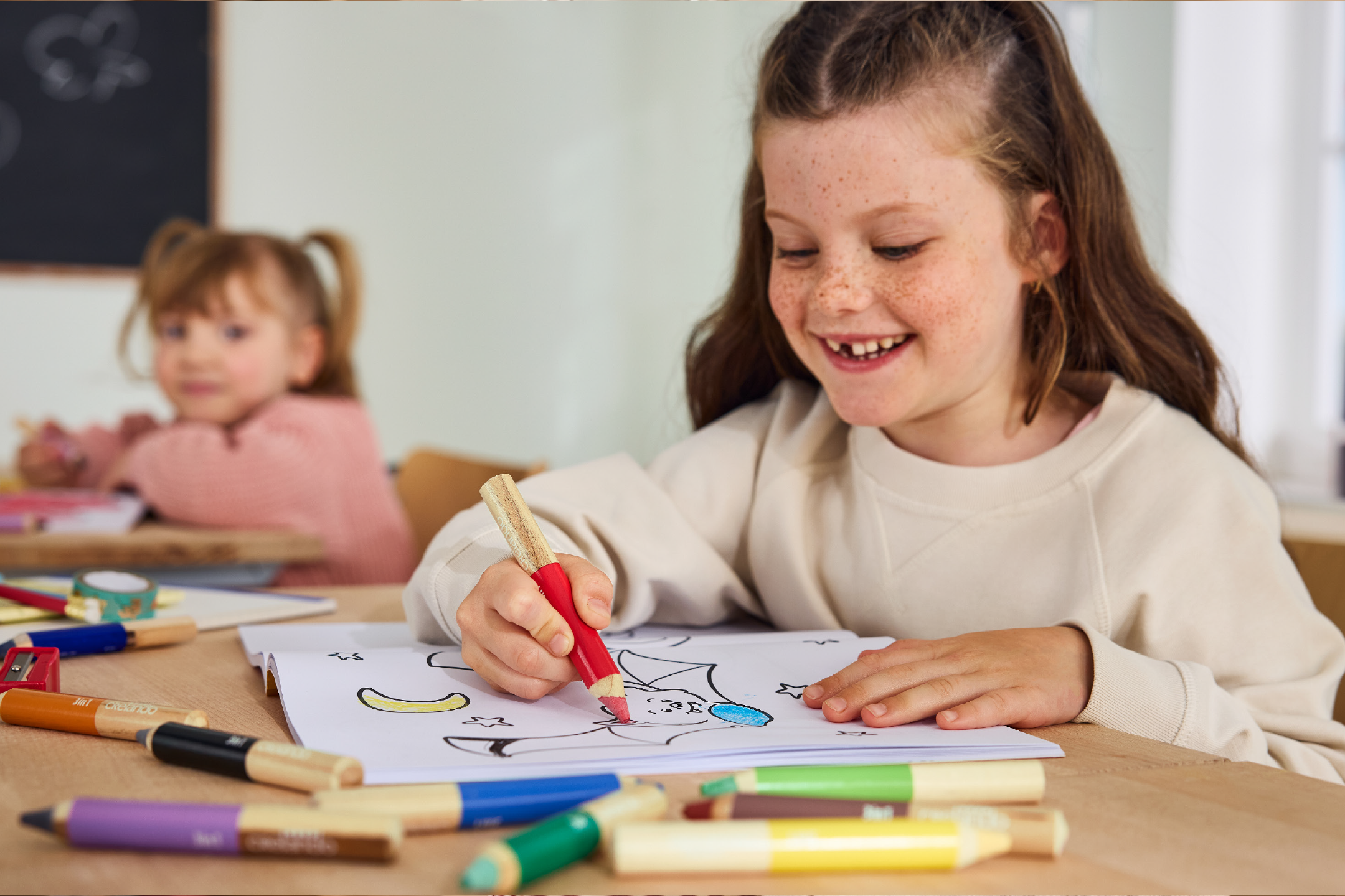 Χαμογελαστή κοπέλα χρωματίζει με ξυλομπογιές Crelando, ένα μικρό παιδί στο βάθος.