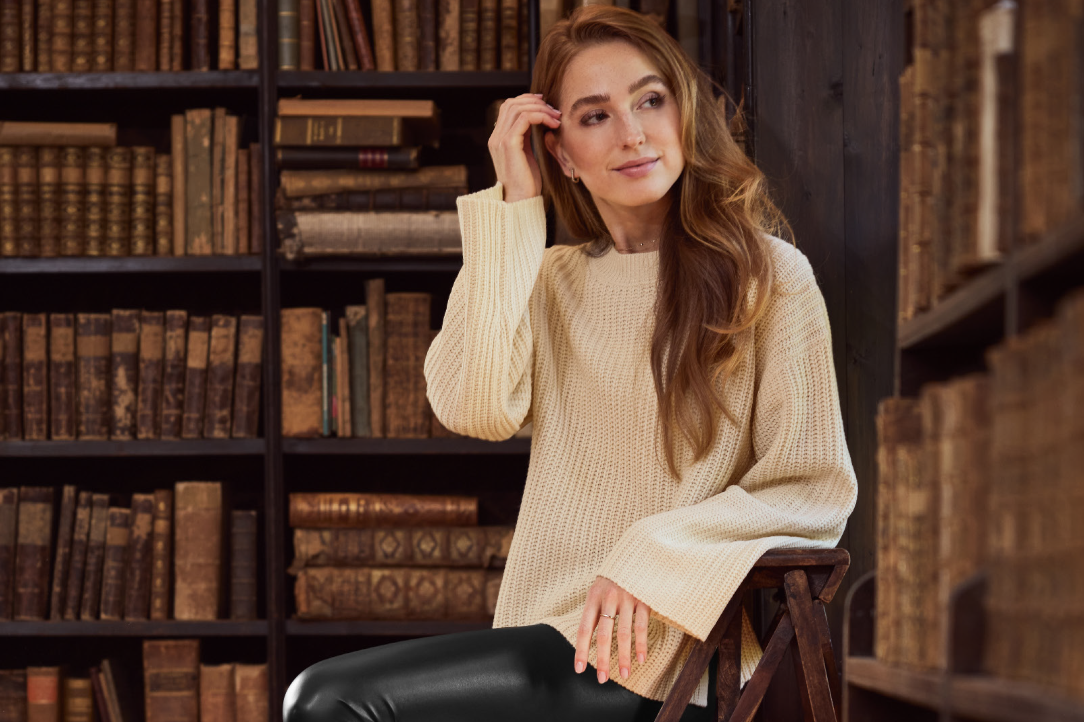 Γυναίκα με κρεμ πουλόβερ και μαύρο παντελόνι σε βιβλιοθήκη.