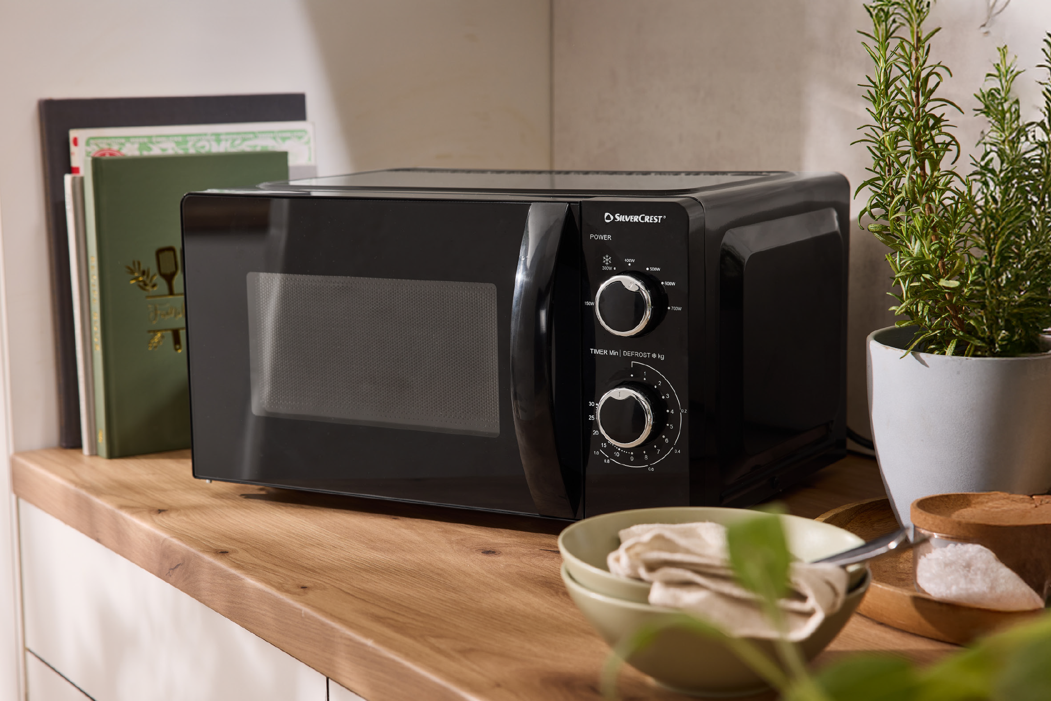 Μαύρος φούρνος μικροκυμάτων σε ξύλινο πάγκο κουζίνας με βιβλία και φυτά.