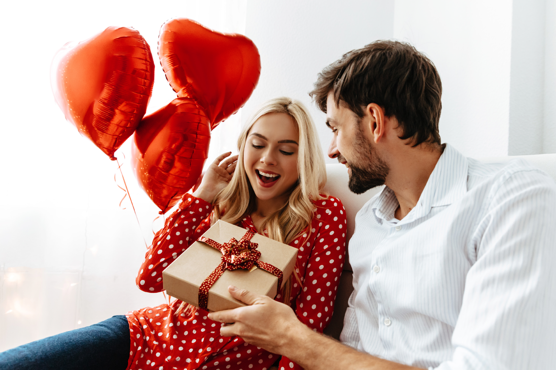 Ένας άντρας δίνει ένα δώρο σε μια χαρούμενη γυναίκα, με κόκκινα μπαλόνια σε σχήμα καρδιάς.