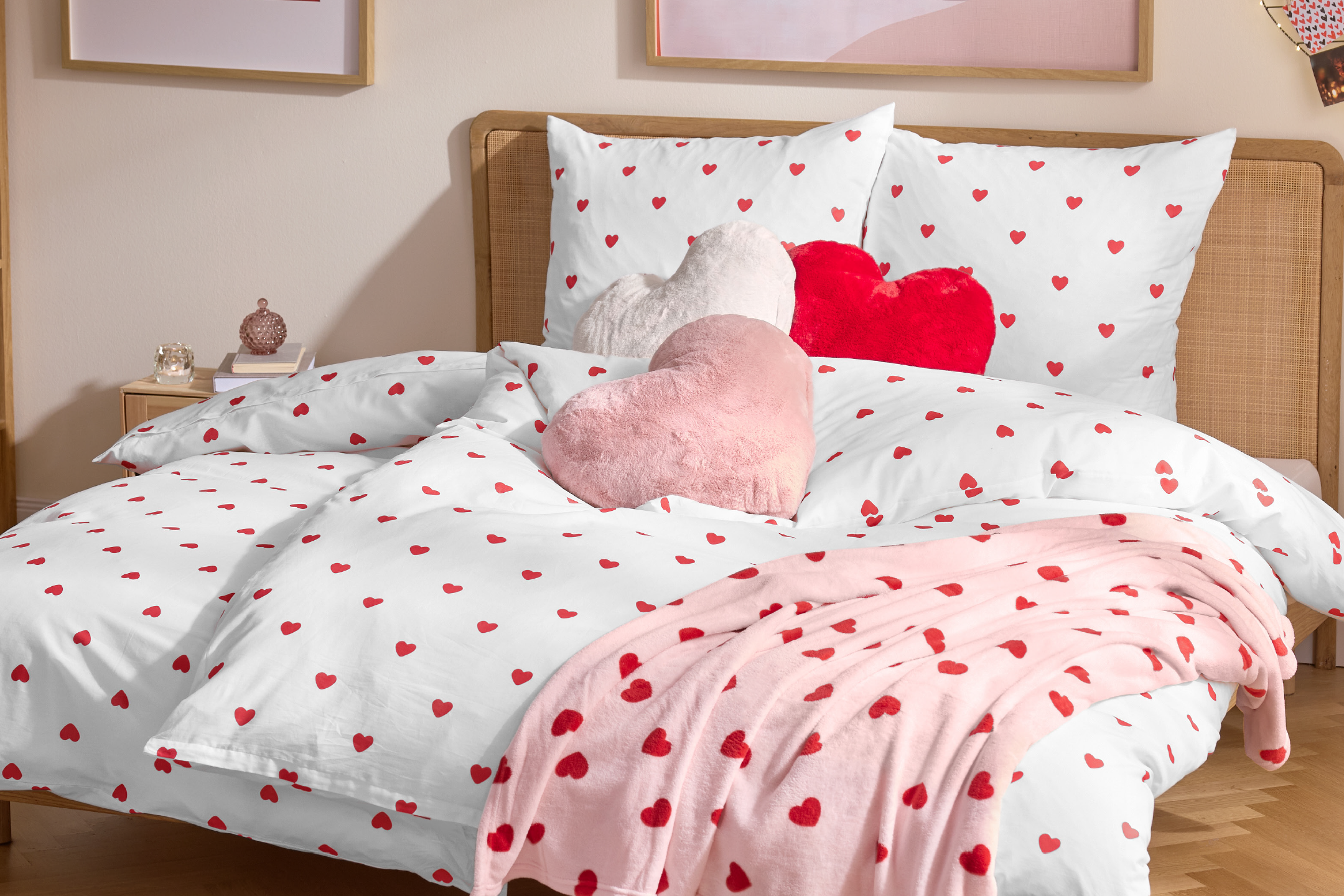 Κρεβάτι με λευκά παπλώματα και μαξιλαροθήκες με κόκκινες καρδιές, διακοσμημένο με τρία βελούδινα μαξιλάρια σε σχήμα καρδιάς και μια ροζ κουβέρτα με καρδιές.
