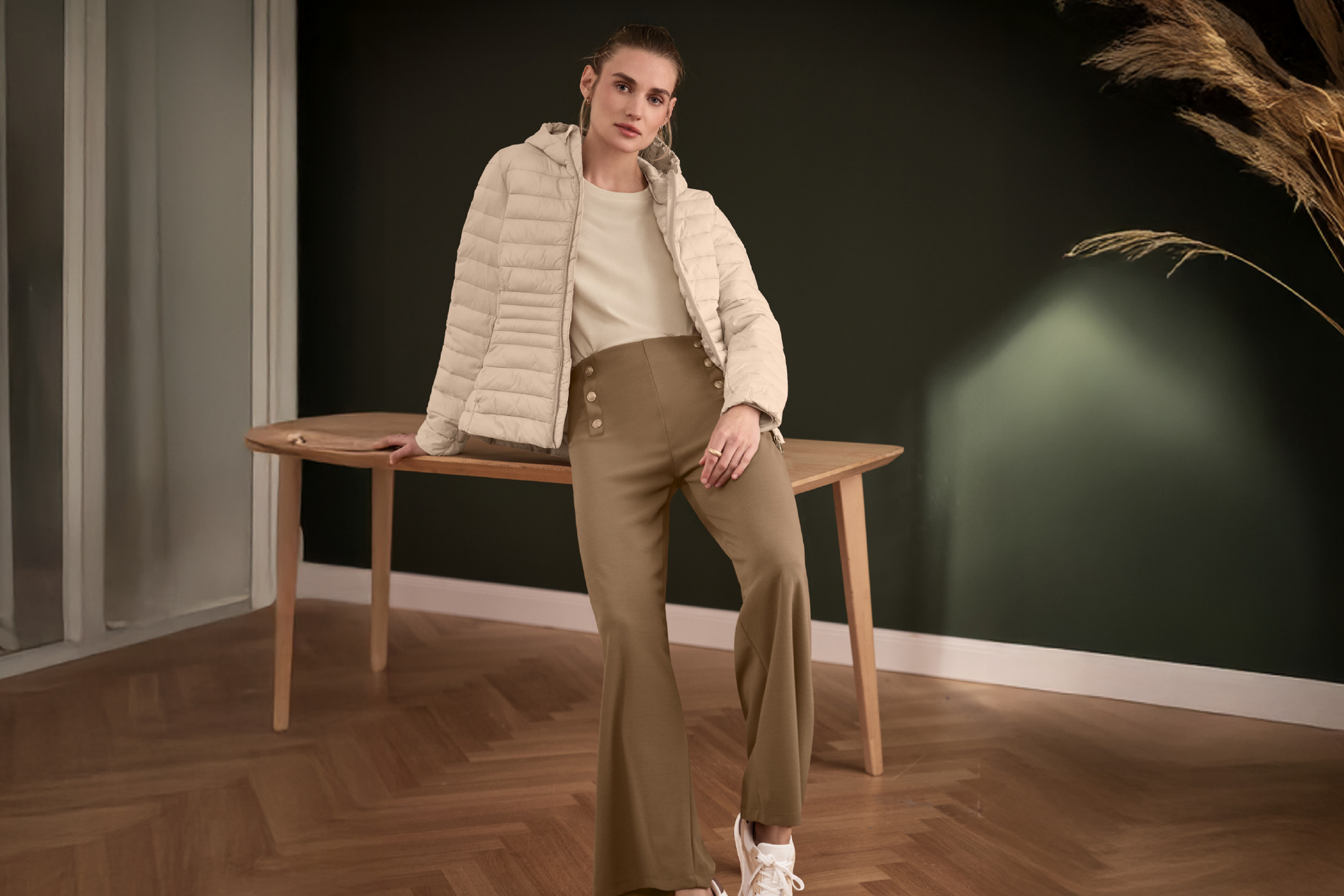 Γυναίκα με μπεζ φουσκωτό μπουφάν, μπεζ μπλούζα και καφέ παντελόνι καμπάνα, ακουμπισμένη σε τραπέζι.