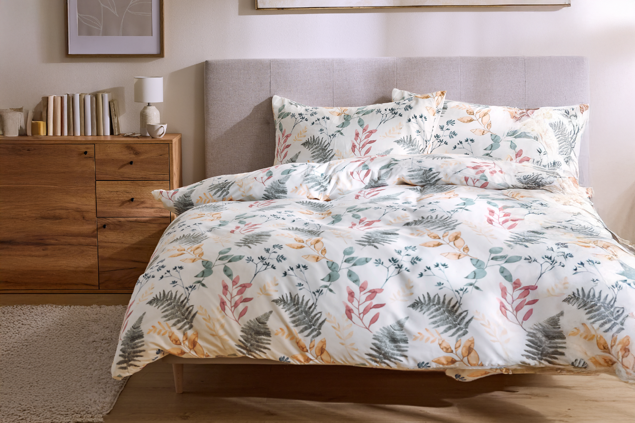 Κρεβάτι με λευκά σεντόνια με σχέδια φύλλων, κομοδίνο με βιβλία και λάμπα.