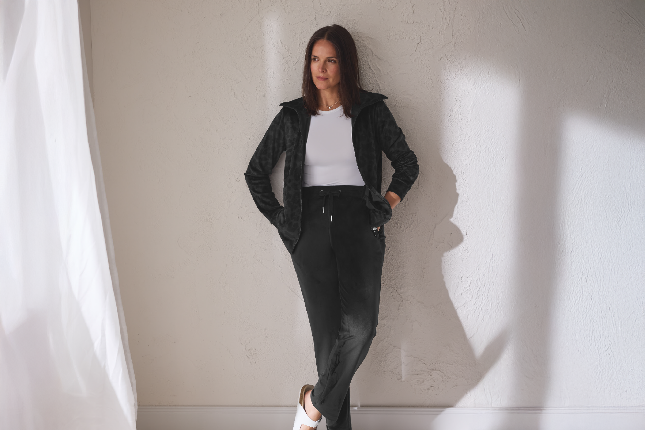 Γυναίκα με μαύρο βελούδινο σετ φόρμας και λευκό τοπ, ακουμπισμένη σε τοίχο.