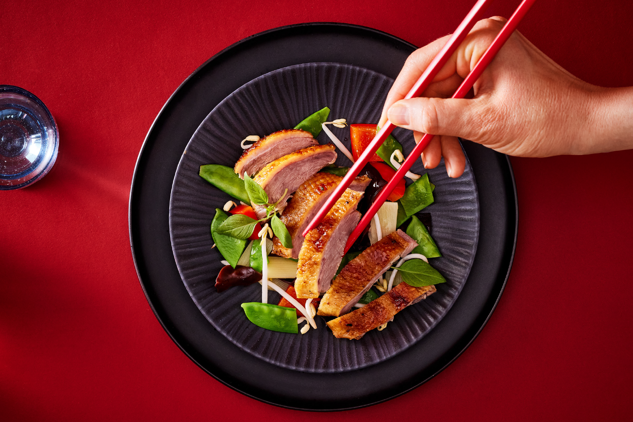 Πάπια με λαχανικά και σάλτσα, σερβιρισμένη σε μαύρο πιάτο, με ένα χέρι να κρατά ξυλάκια.