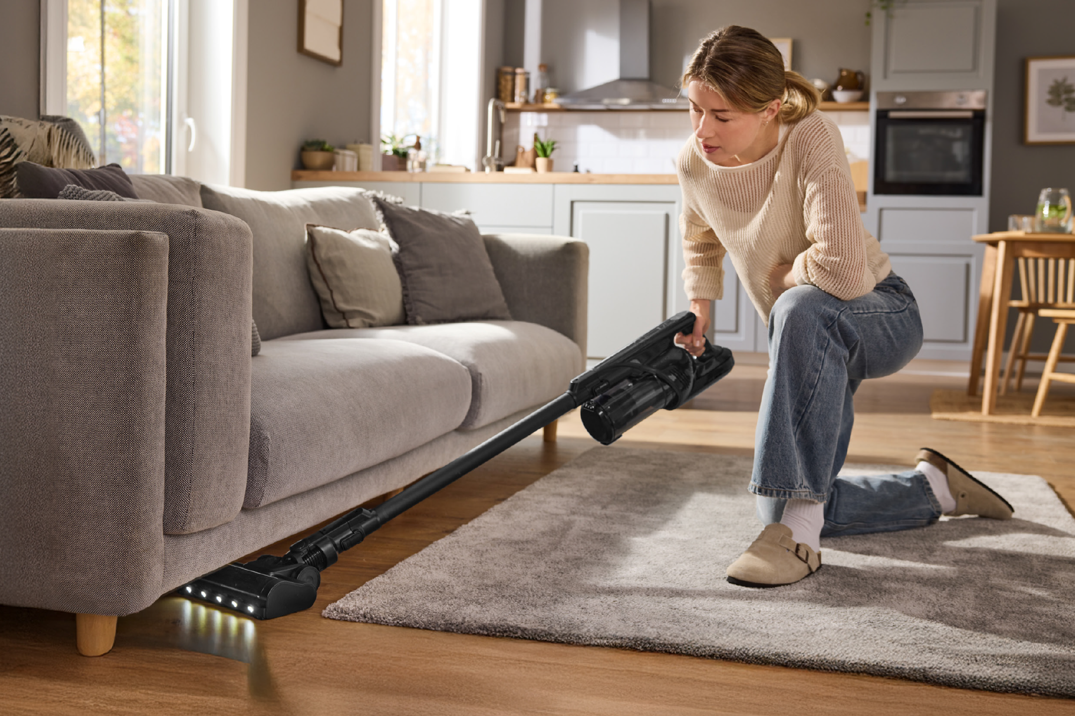 Γυναίκα σκουπίζει κάτω από έναν καναπέ με μια ασύρματη ηλεκτρική σκούπα.