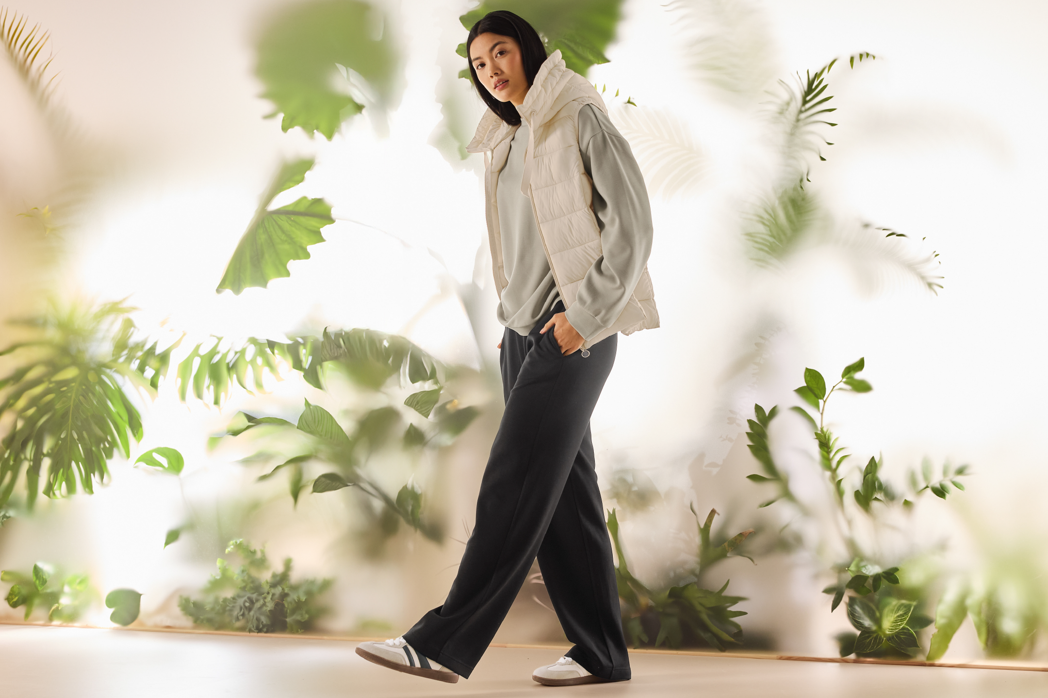 Γυναίκα με μπεζ αμάνικο μπουφάν, γκρι φούτερ και μαύρο παντελόνι φόρμας, ανάμεσα σε φυτά.