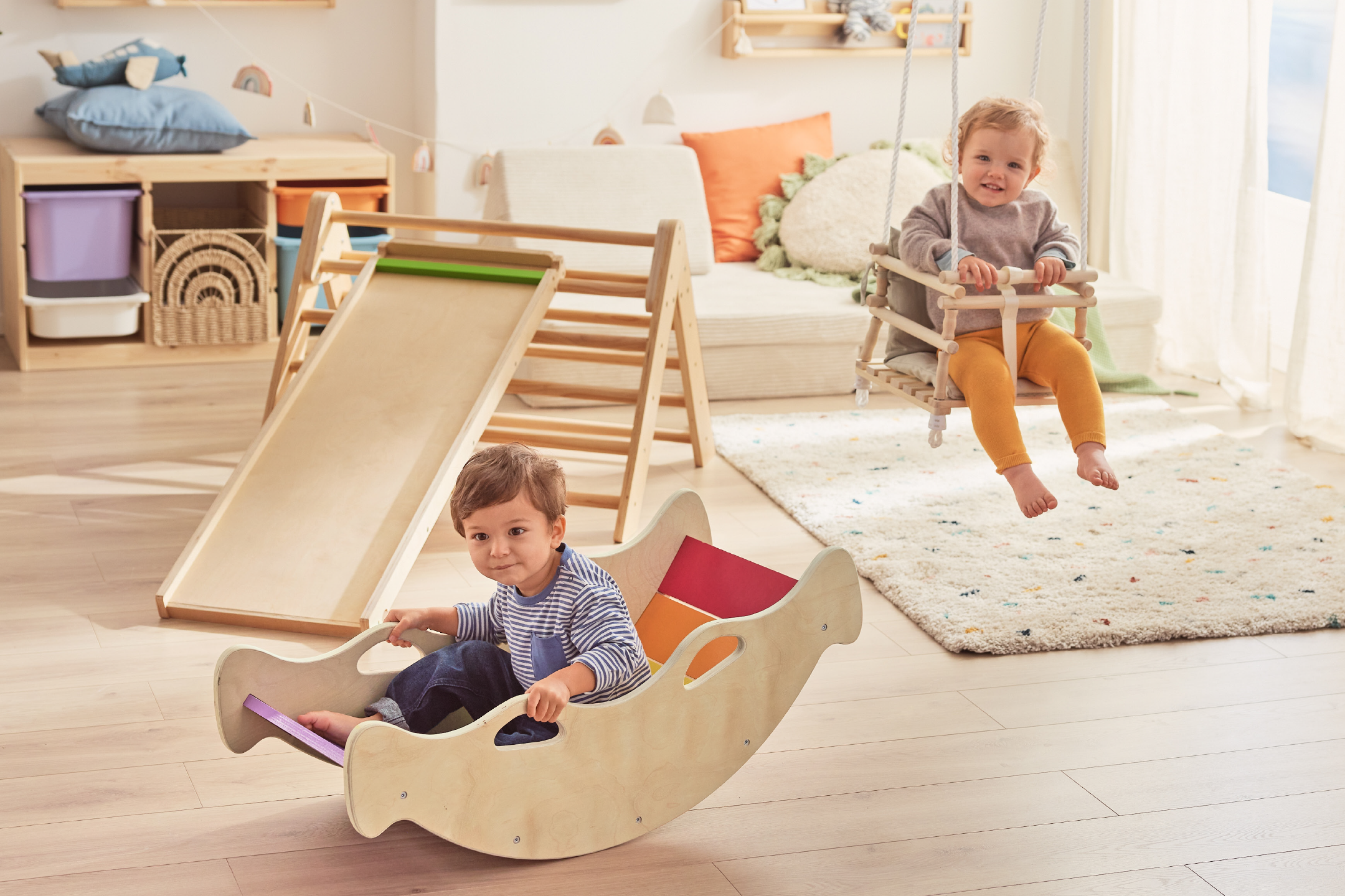 Δύο παιδιά παίζουν σε ένα παιδικό δωμάτιο με ξύλινη τσουλήθρα, κούνια και κουνιστή βάρκα.