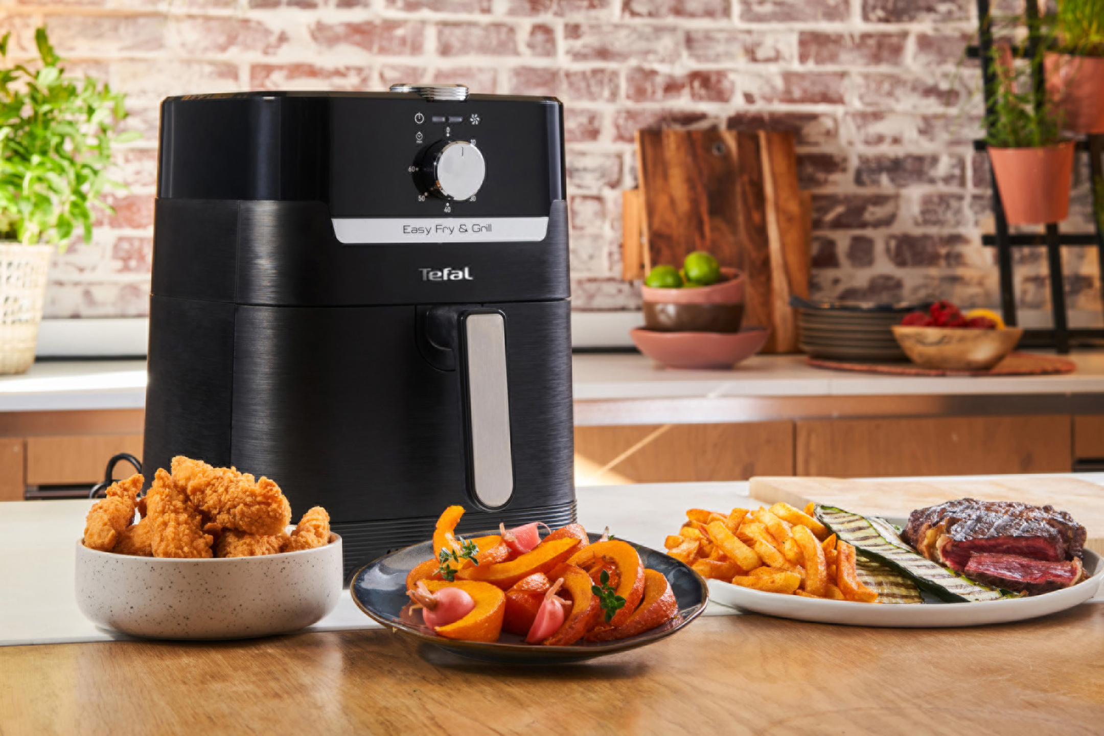 Μαύρη φριτέζα αέρος και ψησταριά σε πάγκο κουζίνας με τηγανητό κοτόπουλο, λαχανικά και μπριζόλα.