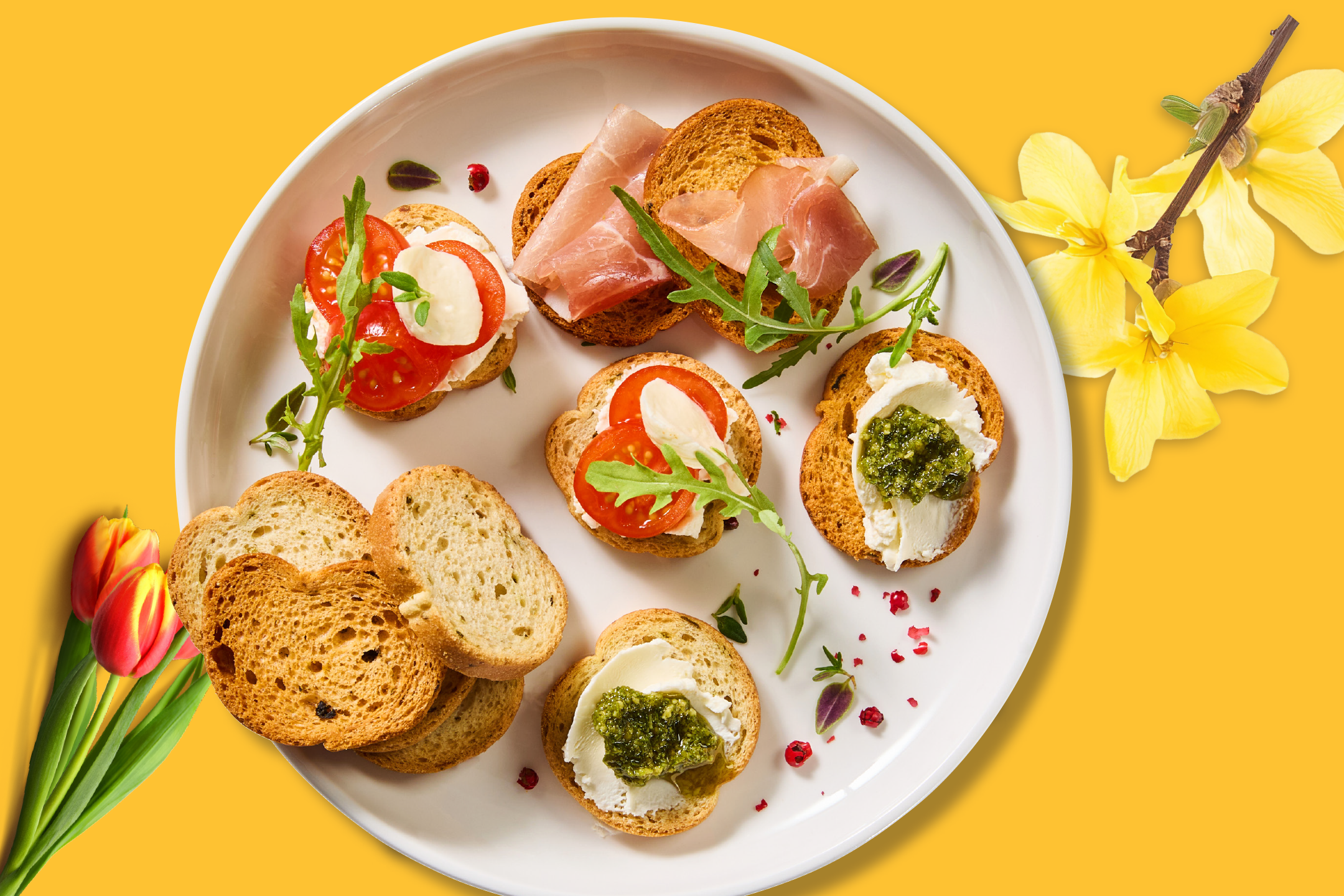 Ποικιλία από μπρουσκέτες με ντομάτα, μοτσαρέλα, προσούτο και πέστο, σερβιρισμένες σε λευκό πιάτο.