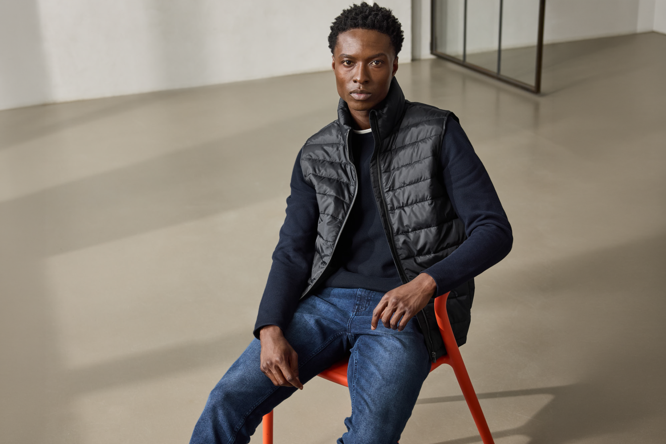 Άνδρας φοράει μαύρο αμάνικο μπουφάν, σκούρο μπλε πουλόβερ και τζιν, καθισμένος σε πορτοκαλί καρέκλα.