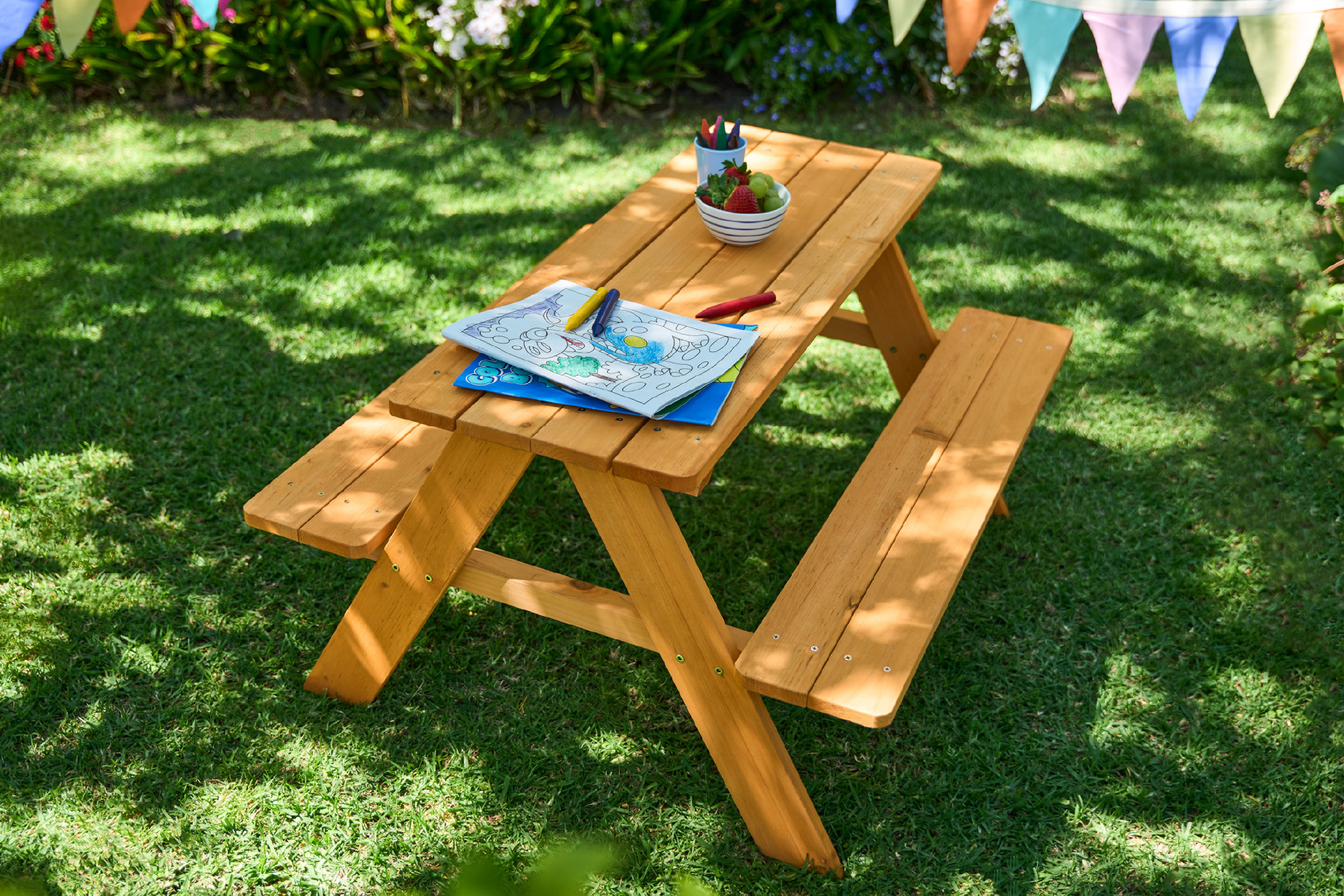Ξύλινο παιδικό τραπέζι πικνίκ με βιβλίο ζωγραφικής, κηρομπογιές και μπολ με φρούτα σε κήπο.