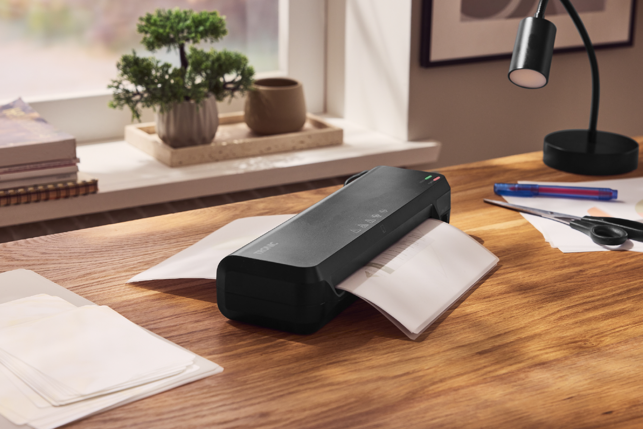 Μαύρη συσκευή πλαστικοποίησης σε ξύλινο γραφείο με χαρτιά, ψαλίδι και επιτραπέζιο φωτιστικό.