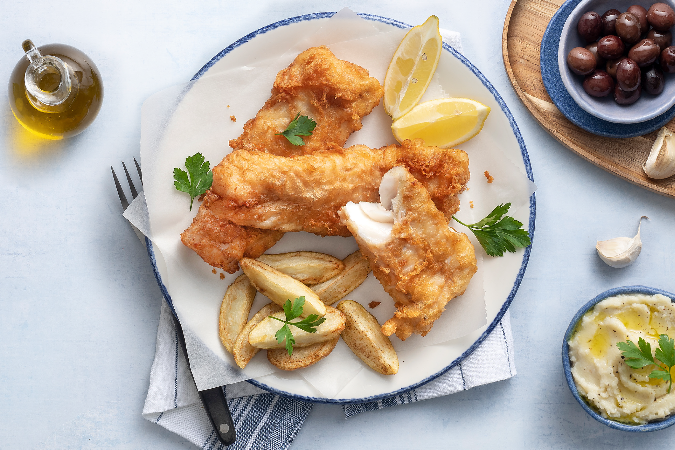 Τηγανητό ψάρι με πατάτες, λεμόνι, ελιές και σκορδαλιά σε λευκό πιάτο.