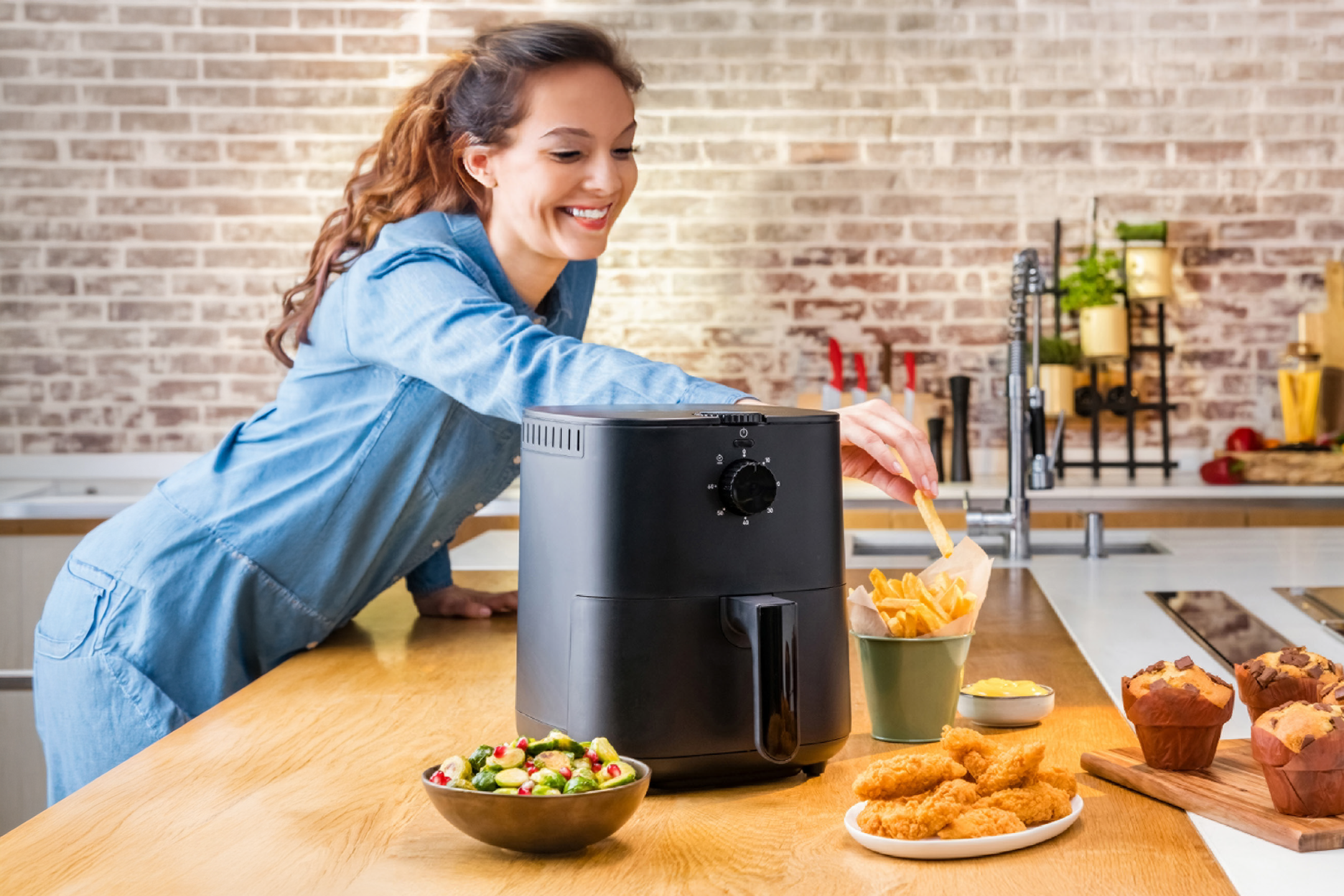 Γυναίκα χρησιμοποιεί φριτέζα αέρος για να φτιάξει τηγανητές πατάτες, κοτομπουκιές και σαλάτα.