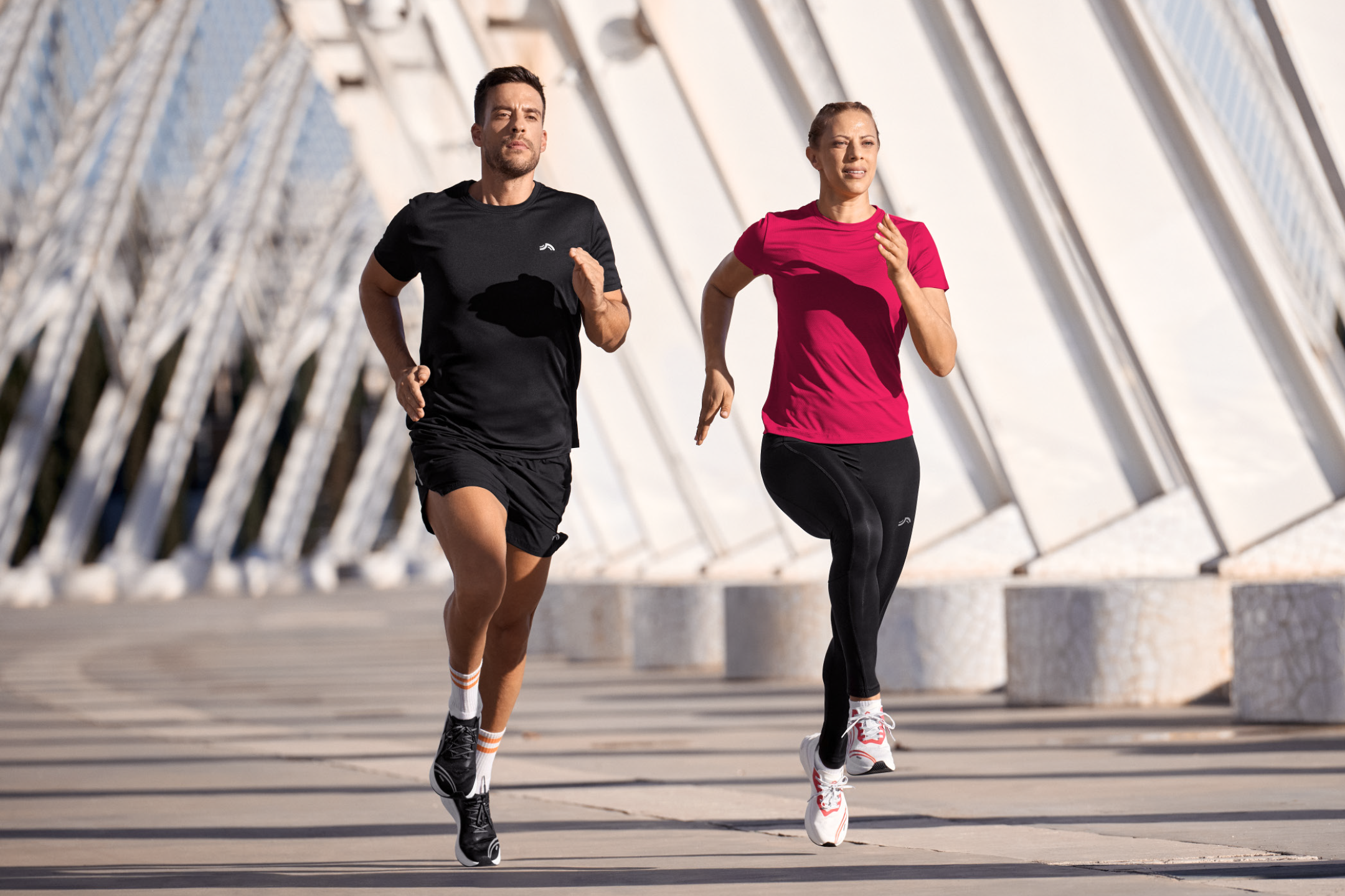 Δύο δρομείς με αθλητικά ρούχα τρέχουν σε εξωτερικό χώρο με μοντέρνα αρχιτεκτονική στο παρασκήνιο.