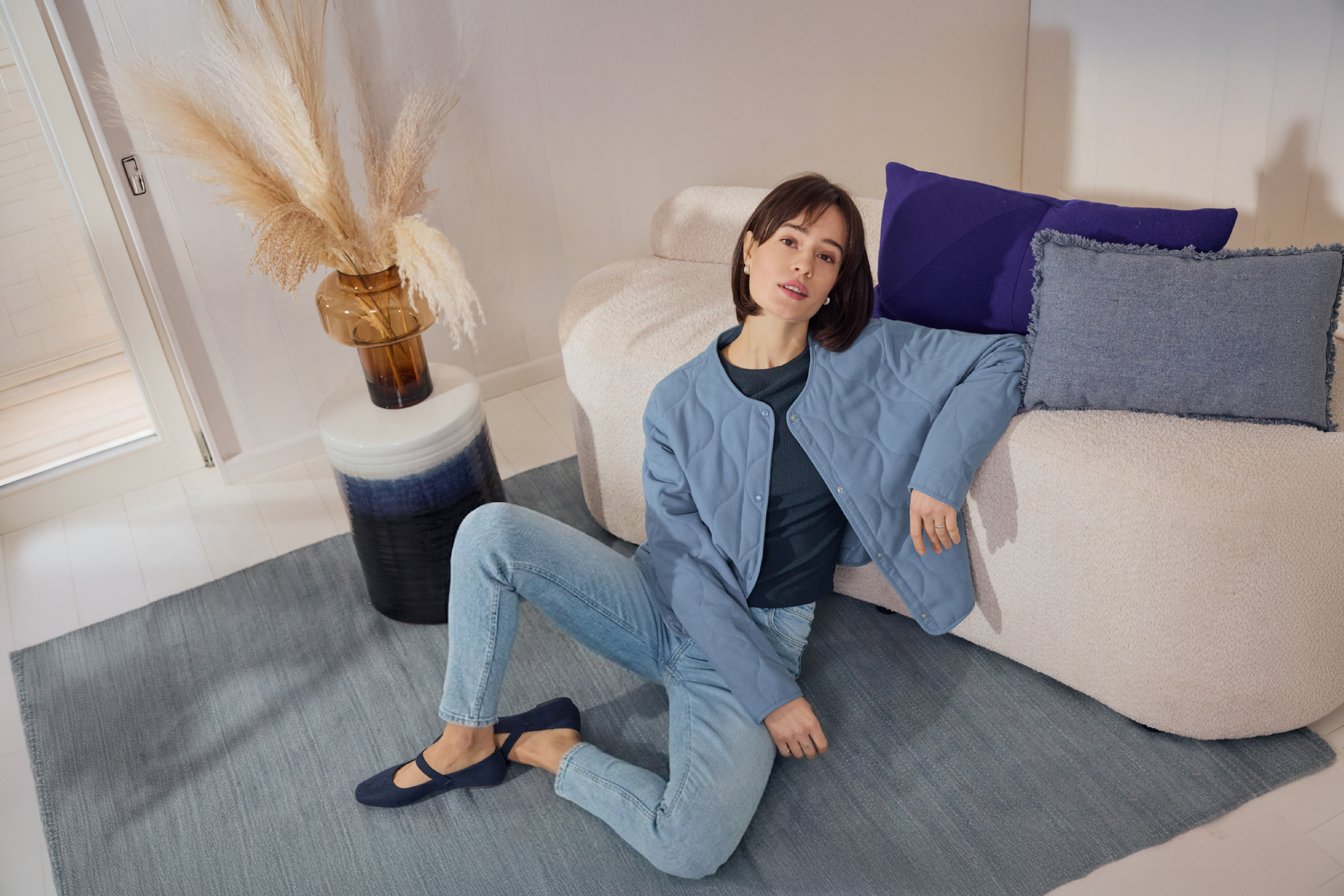 Γυναίκα με μπλε καπιτονέ μπουφάν και τζιν, καθισμένη σε χαλί δίπλα σε μπεζ καναπέ με μαξιλάρια.