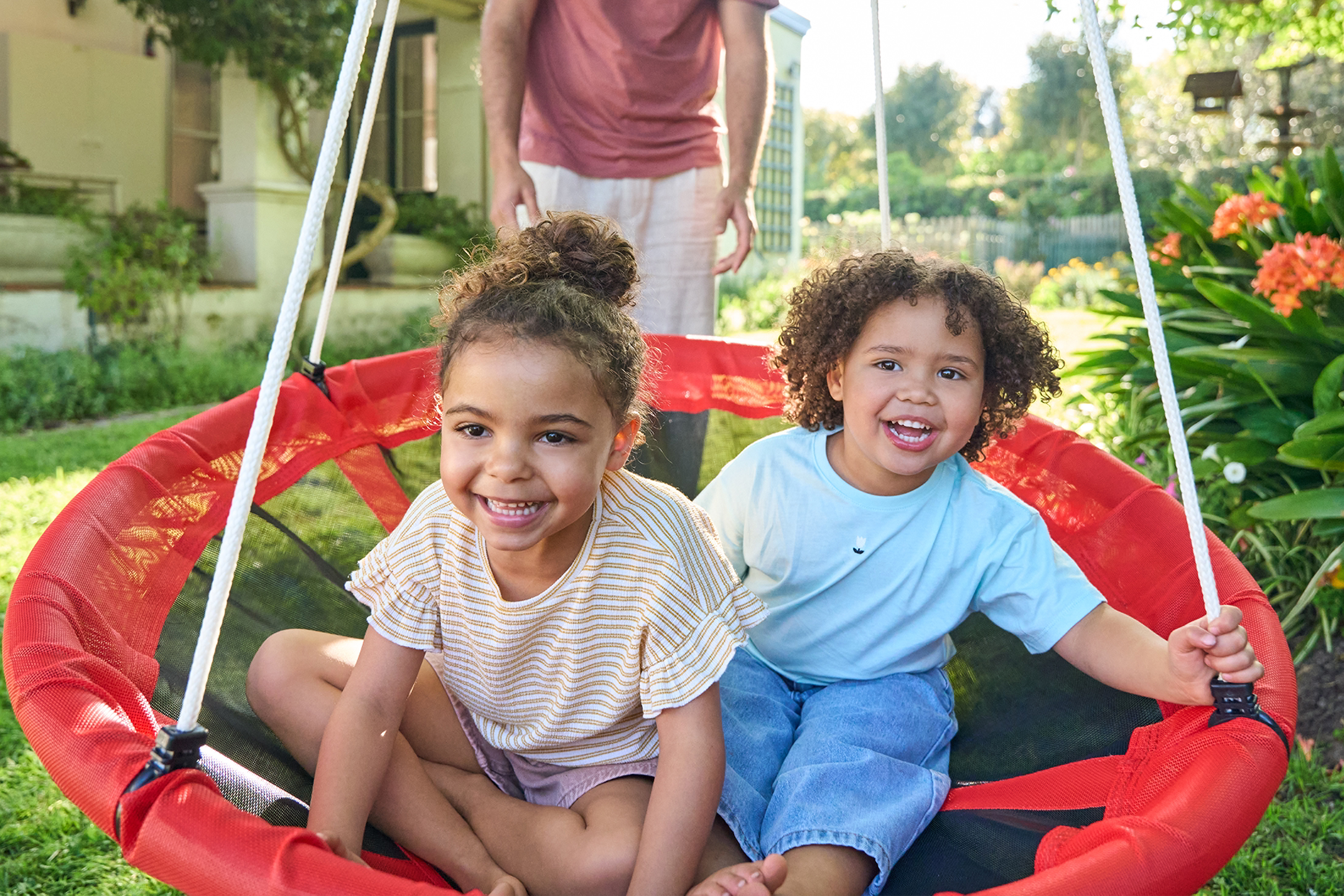 Δύο παιδιά σε μια κόκκινη κούνια, με έναν ενήλικα να στέκεται από πίσω.