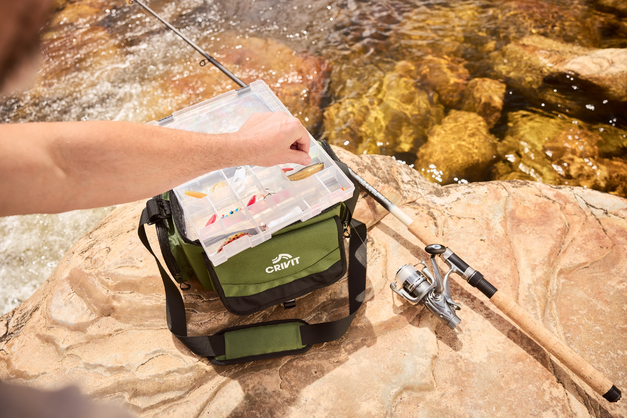 Άντρας ψαρεύει, επιλέγοντας δόλωμα από κουτί με αλιευτικά είδη, δίπλα σε καλάμι ψαρέματος και τσάντα.