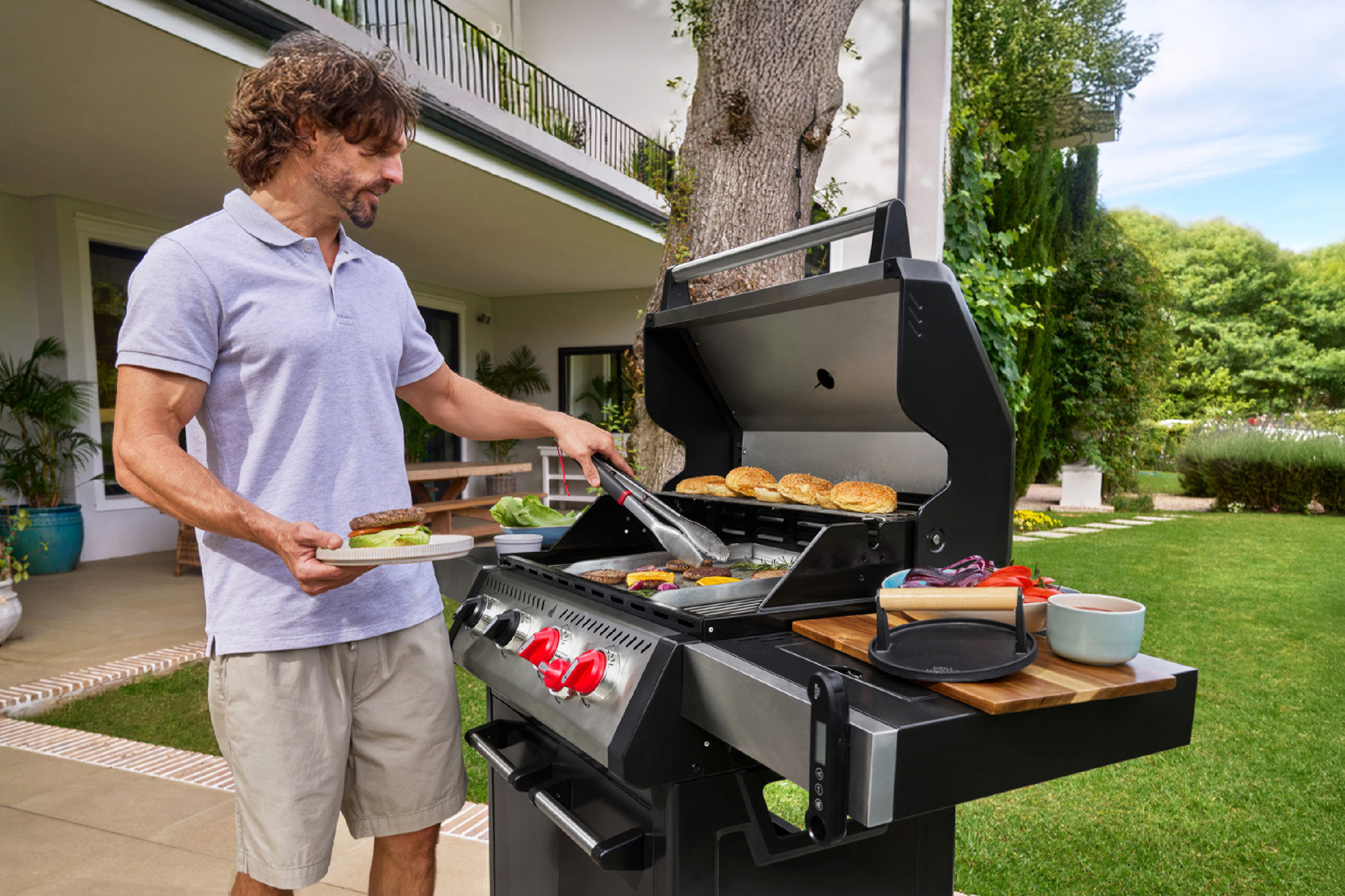 Άντρας ψήνει μπιφτέκια και ψωμάκια για μπέργκερ σε ψησταριά υγραερίου στον κήπο.