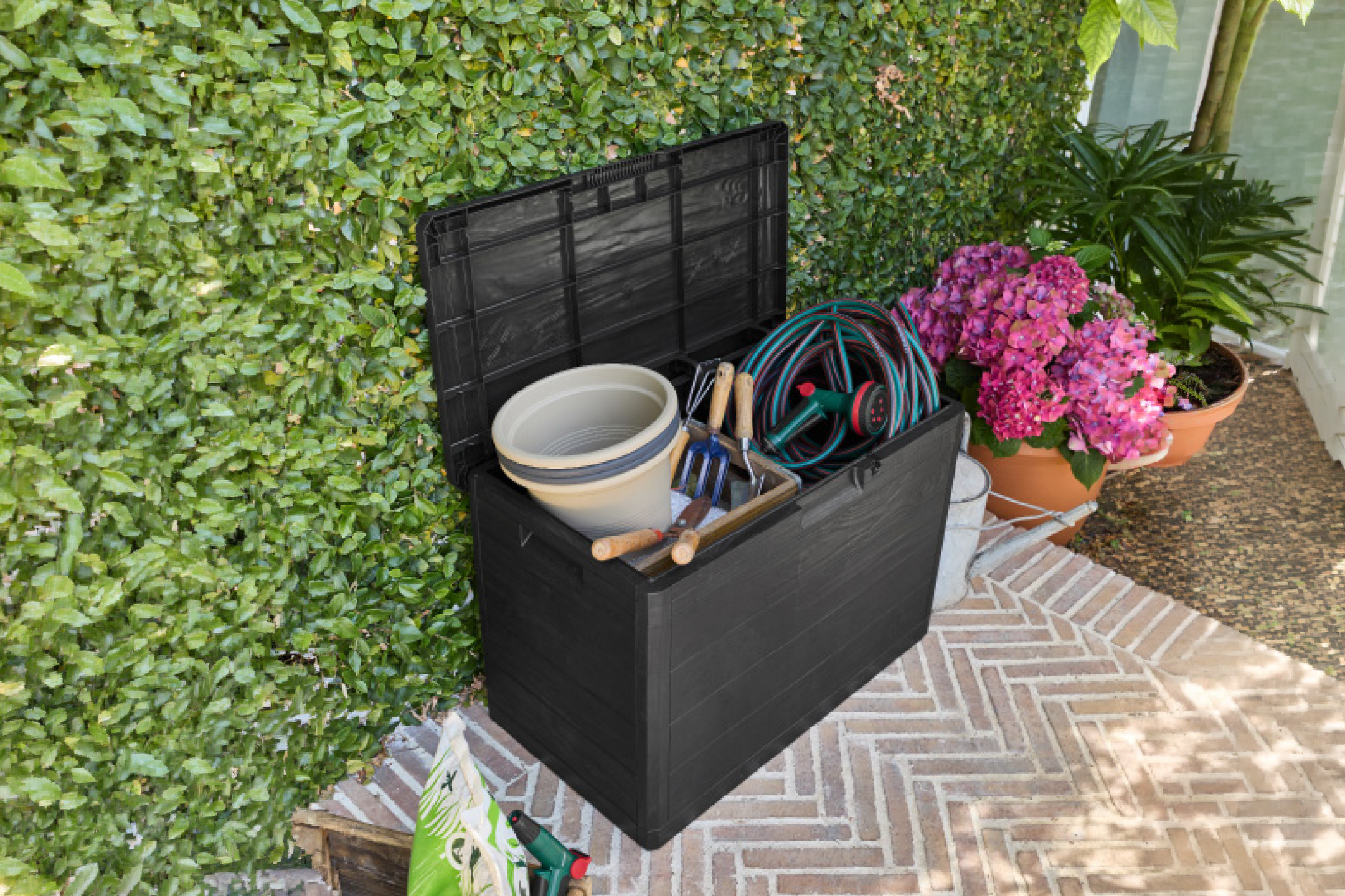 Μαύρο κουτί αποθήκευσης κήπου γεμάτο με εργαλεία κηπουρικής και λάστιχο ποτίσματος, δίπλα σε φυτά.