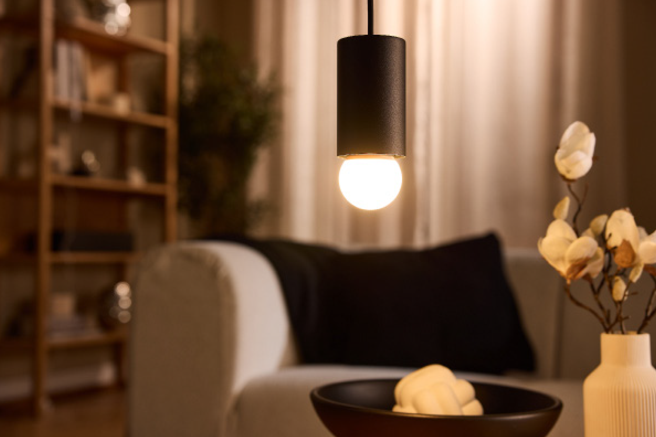 Κρεμαστό φωτιστικό με ζεστό φως σε μοντέρνο σαλόνι με καναπέ και ράφια.