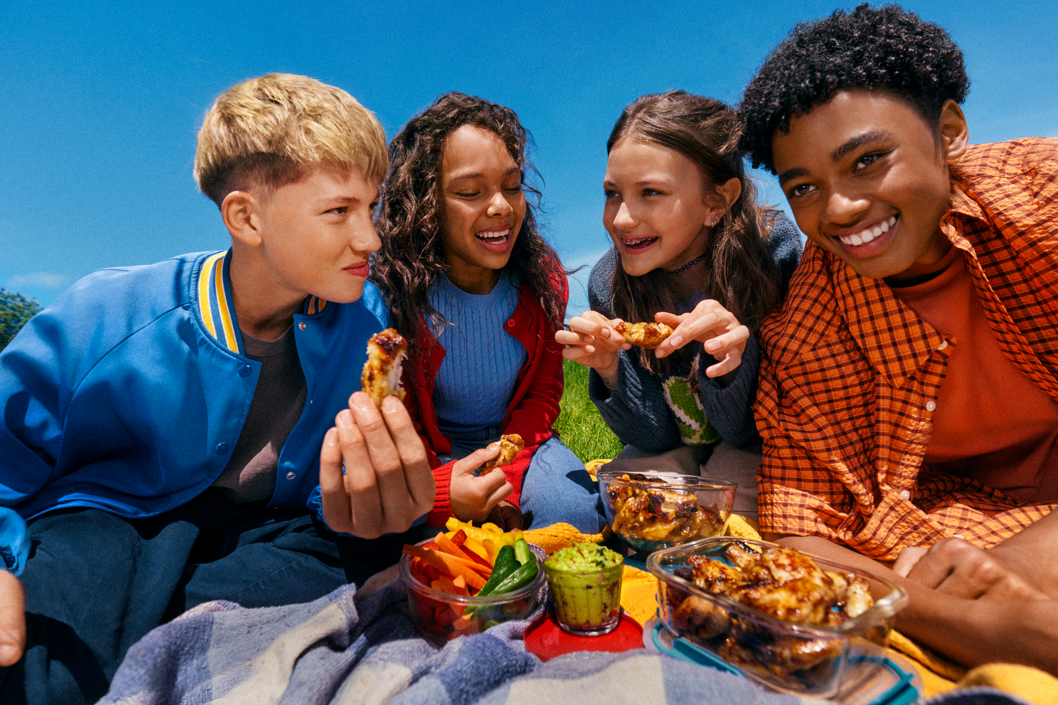 Παιδιά σε πικνίκ τρώνε φτερούγες κοτόπουλου, λαχανικά και γουακαμόλε.