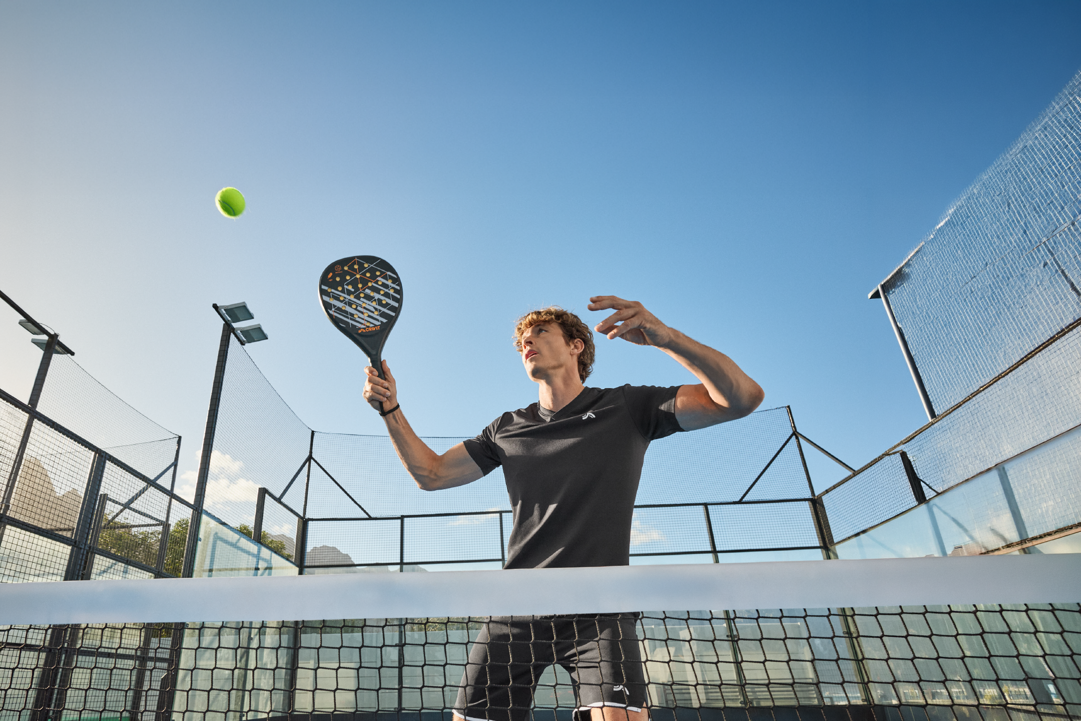 Άνδρας παίζει padel σε εξωτερικό γήπεδο, κρατώντας ρακέτα και χτυπώντας μπάλα.