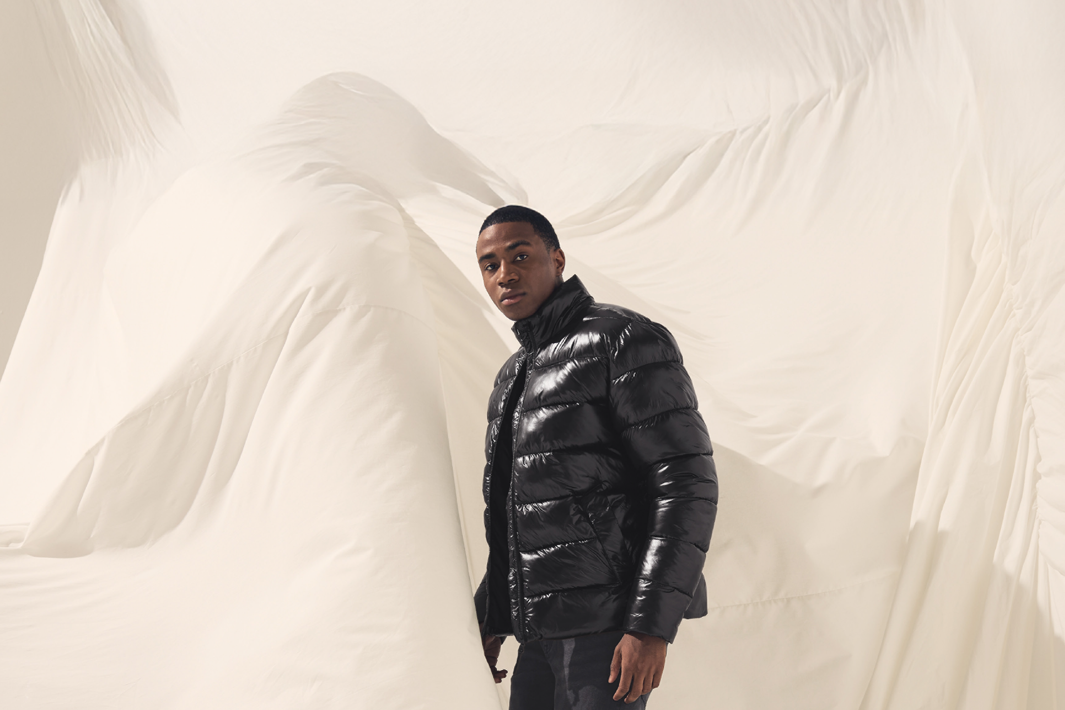 Ένας άντρας φοράει ένα μαύρο γυαλιστερό φουσκωτό μπουφάν μπροστά σε ένα λευκό φόντο από ύφασμα.