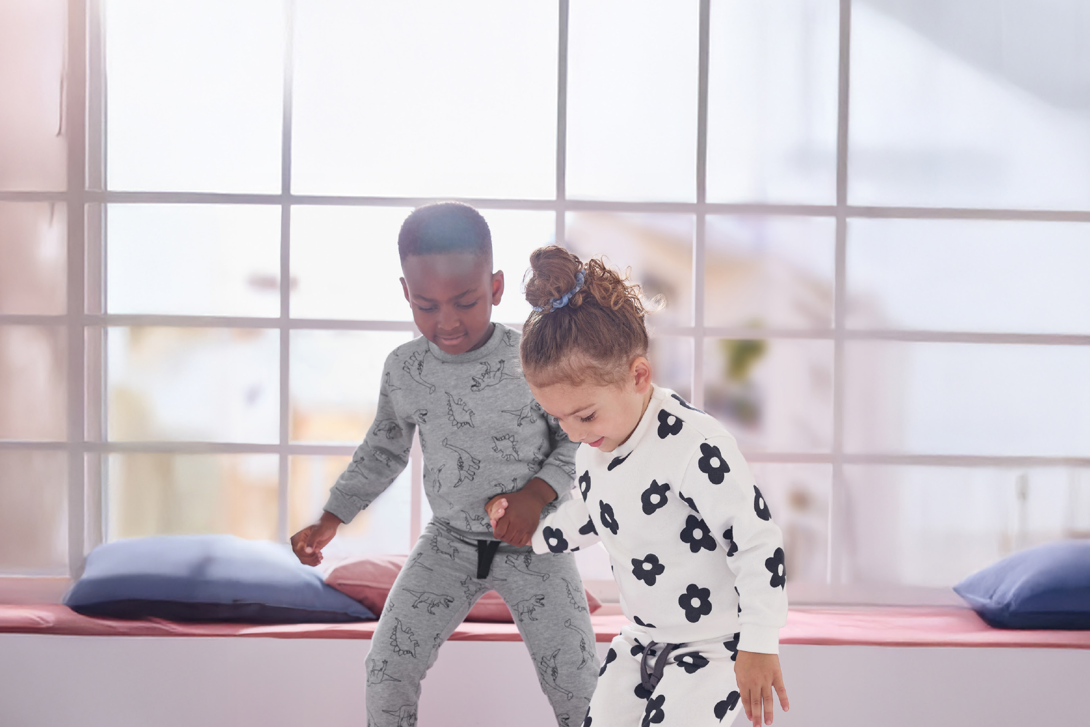 Παιδιά με πιτζάμες με σχέδια δεινοσαύρων και λουλουδιών, παίζουν μπροστά σε ένα παράθυρο.