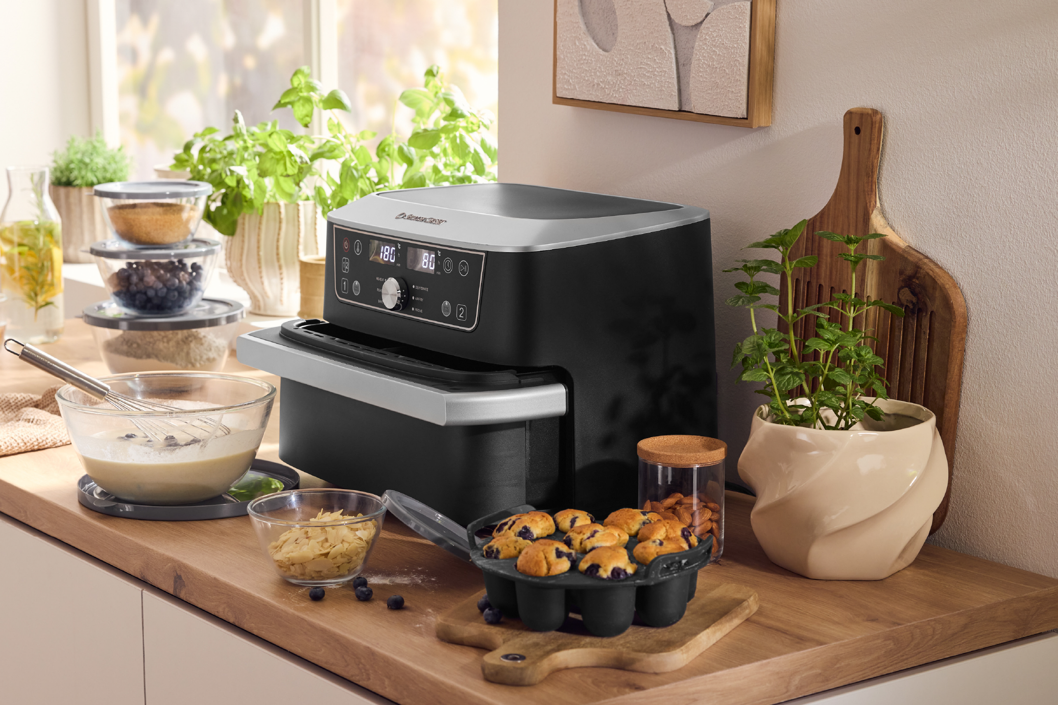 Μαύρη φριτέζα αέρος Silvercrest με ψηφιακή οθόνη, μπολ με υλικά και muffins με μύρτιλα.