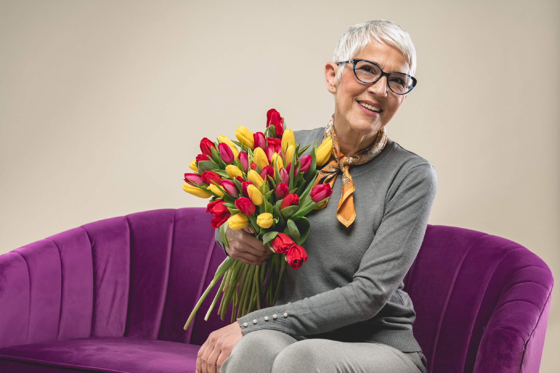 Χαμογελαστή ηλικιωμένη γυναίκα με γυαλιά, κρατώντας μια μεγάλη ανθοδέσμη με κόκκινες και κίτρινες τουλίπες, καθισμένη σε έναν μοβ καναπέ.