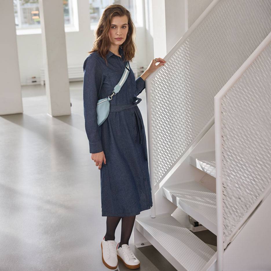 Γυναίκα με τζιν φόρεμα-πουκάμισο, μαύρο καλσόν και λευκά αθλητικά, με γαλάζιο τσαντάκι μέσης.